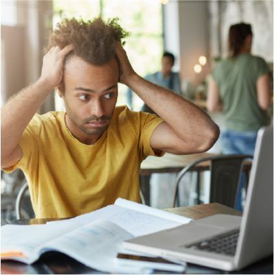 Man with curly hair holds his head, looking stressed, at a desk with a laptop and open book.