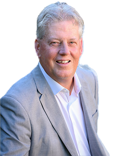Man in light gray suit smiles, leaning forward. White shirt, natural background.