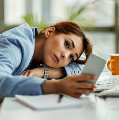 Woman in blue blazer, head resting on desk, looking at phone with a bored expression.