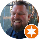 Man with a beard smiling, wearing a suit jacket and blue shirt, with an orange star icon in the bottom corner.