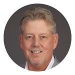 Smiling man in a light gray blazer and a white shirt. The background is gray.