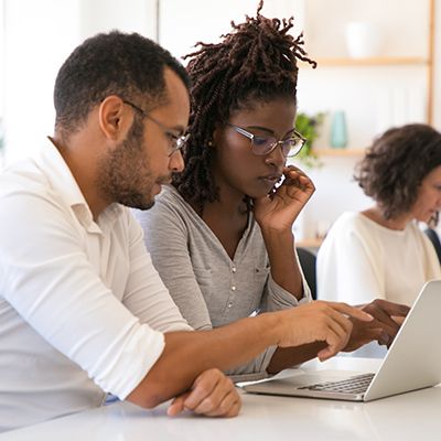 Two people looking at a laptop, one pointing. Another person sits in background.