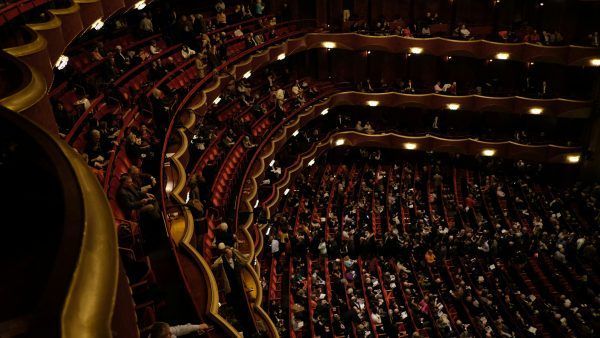 Audience seated in tiered rows of a grand theater, dark red seating, gold accents, dim lighting.