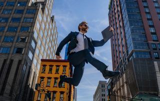 Man in suit jumping with a laptop, between tall buildings on a sunny day.