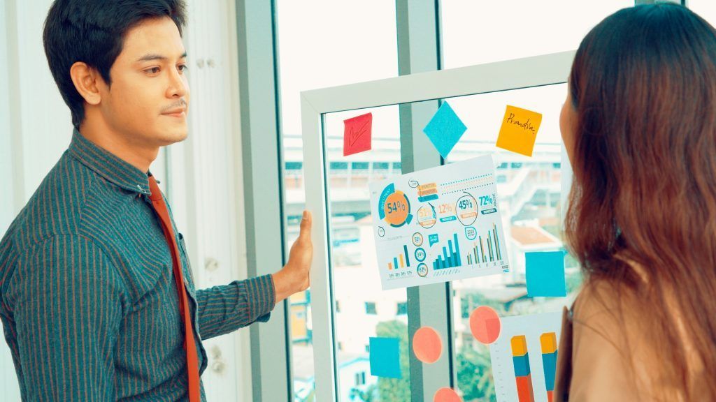 Man points at whiteboard with charts and sticky notes, explaining to a woman in an office setting.