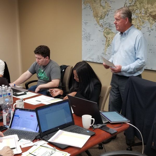 People in a meeting with laptops, papers, and a world map in the background; a man stands presenting.