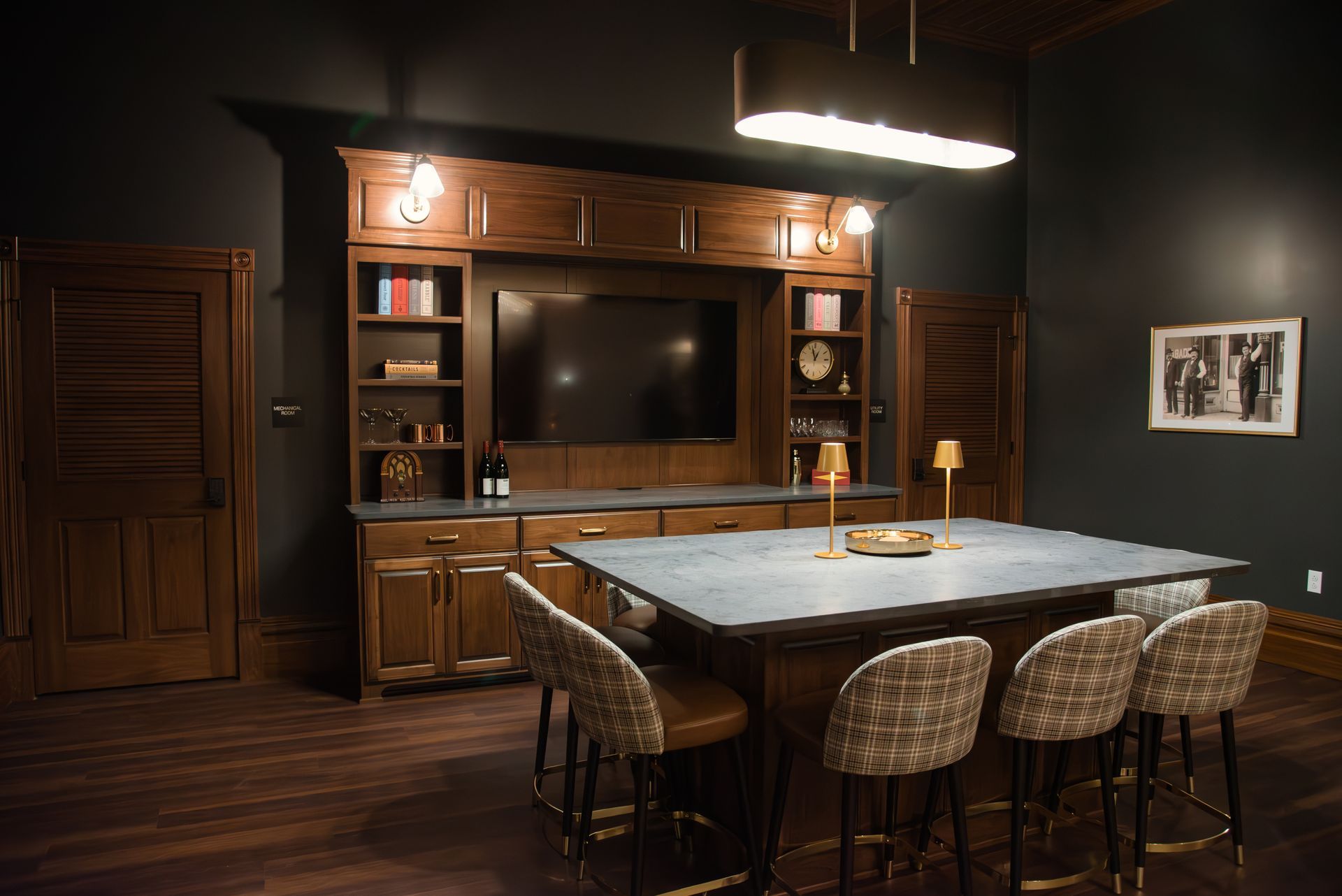 Dark-themed bar area with a built-in wooden cabinet, TV, island table, and bar stools.
