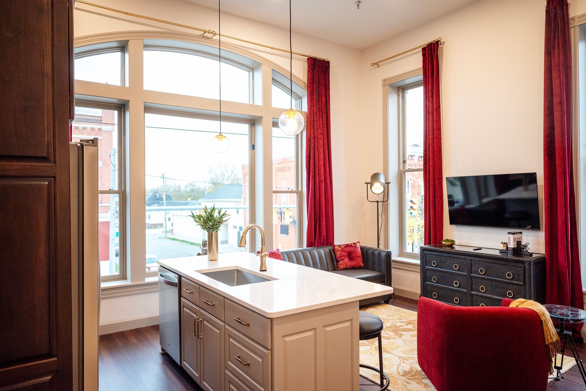 Stylish apartment interior: kitchen island, windows, red curtains, black leather sofa, red armchair, and a TV.