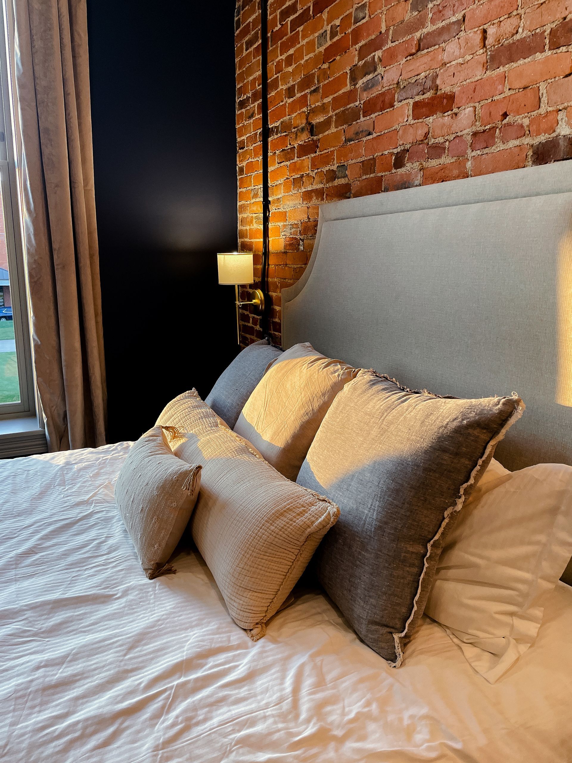 Bed with several pillows in front of a brick wall, next to a dark blue wall and a window.