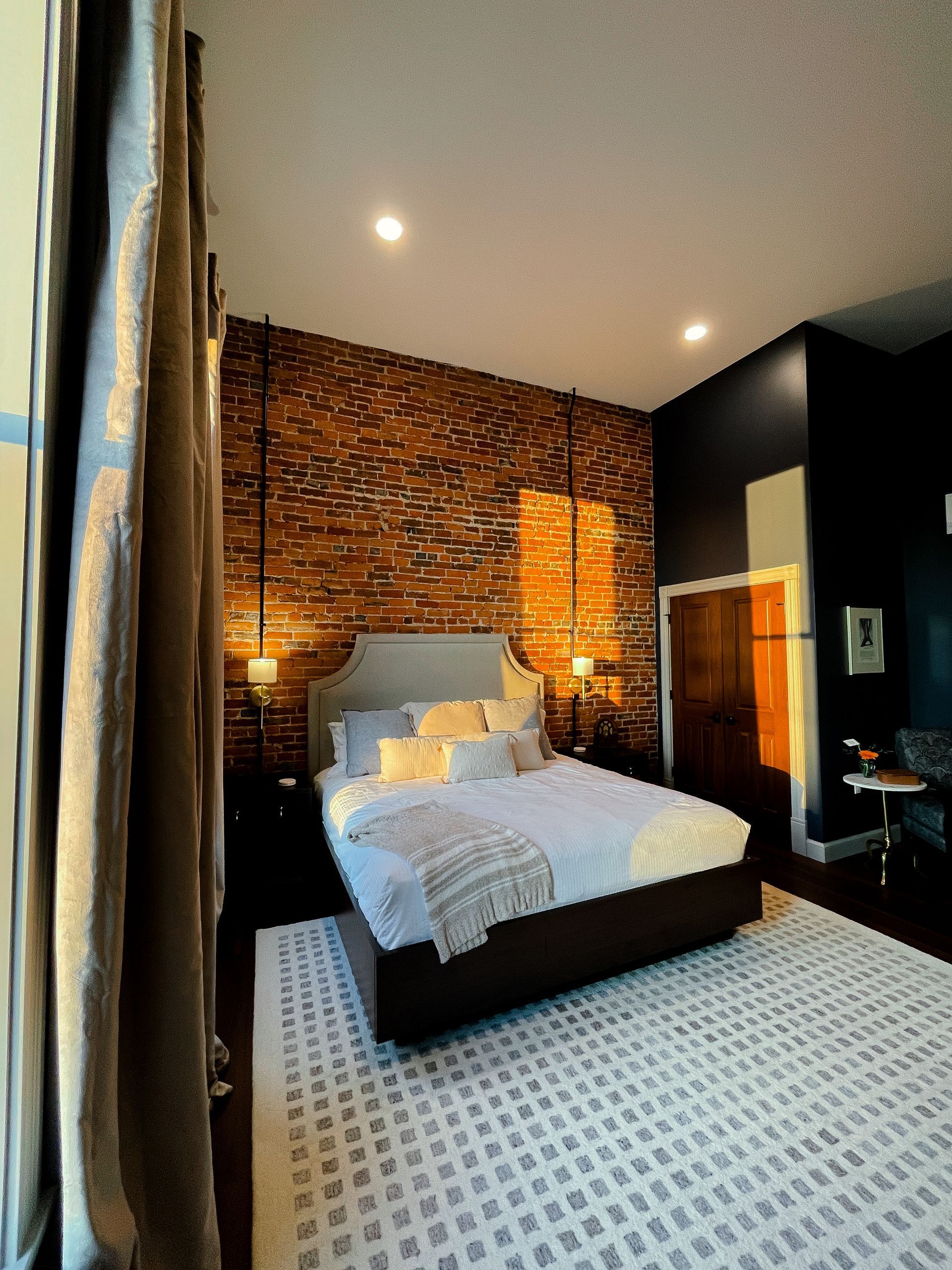 Bedroom with exposed brick wall, white bed, patterned rug, and dark walls.