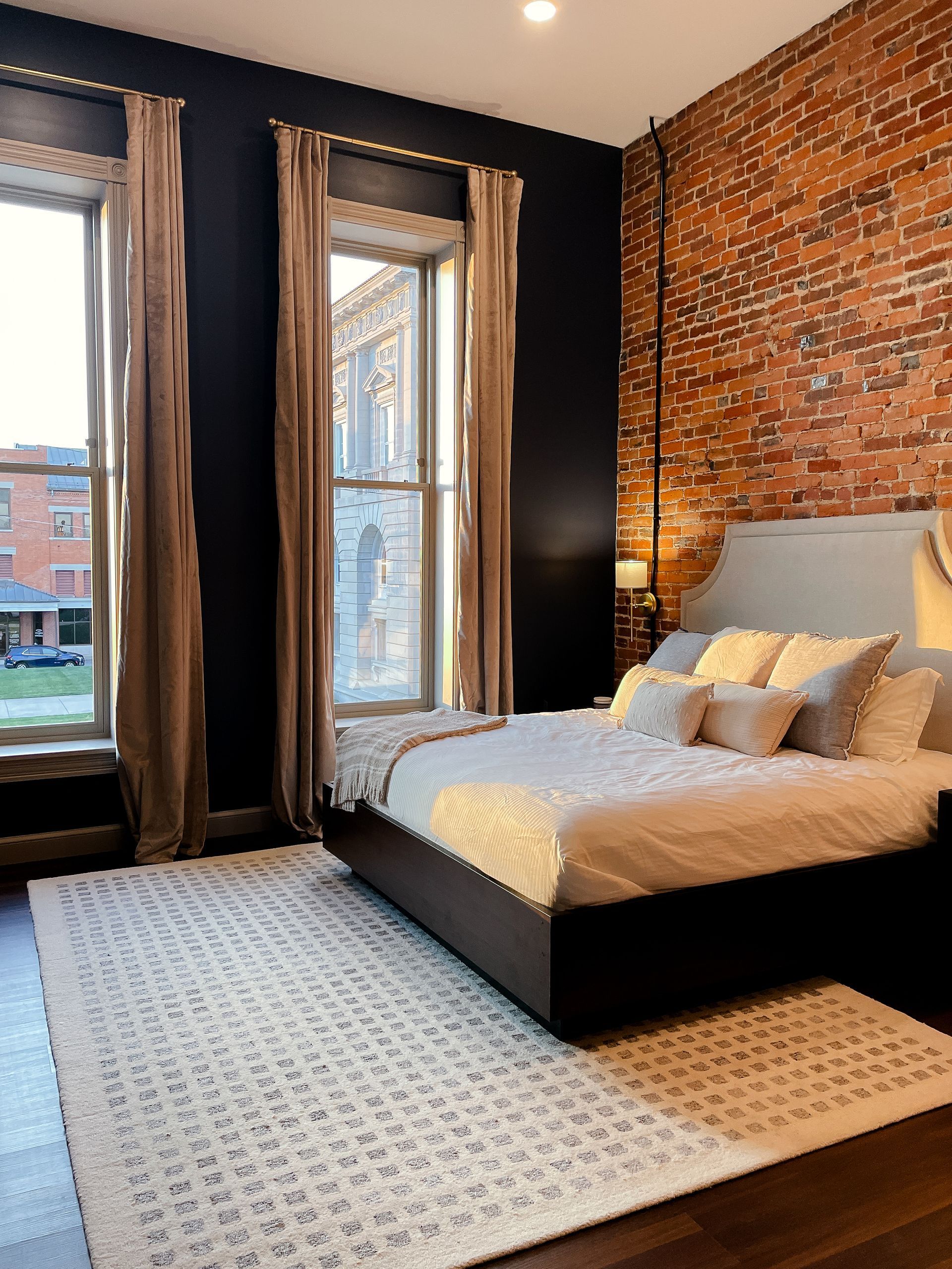 Bedroom with dark walls, exposed brick, and a bed with beige linens. Sunlight streams through the windows.