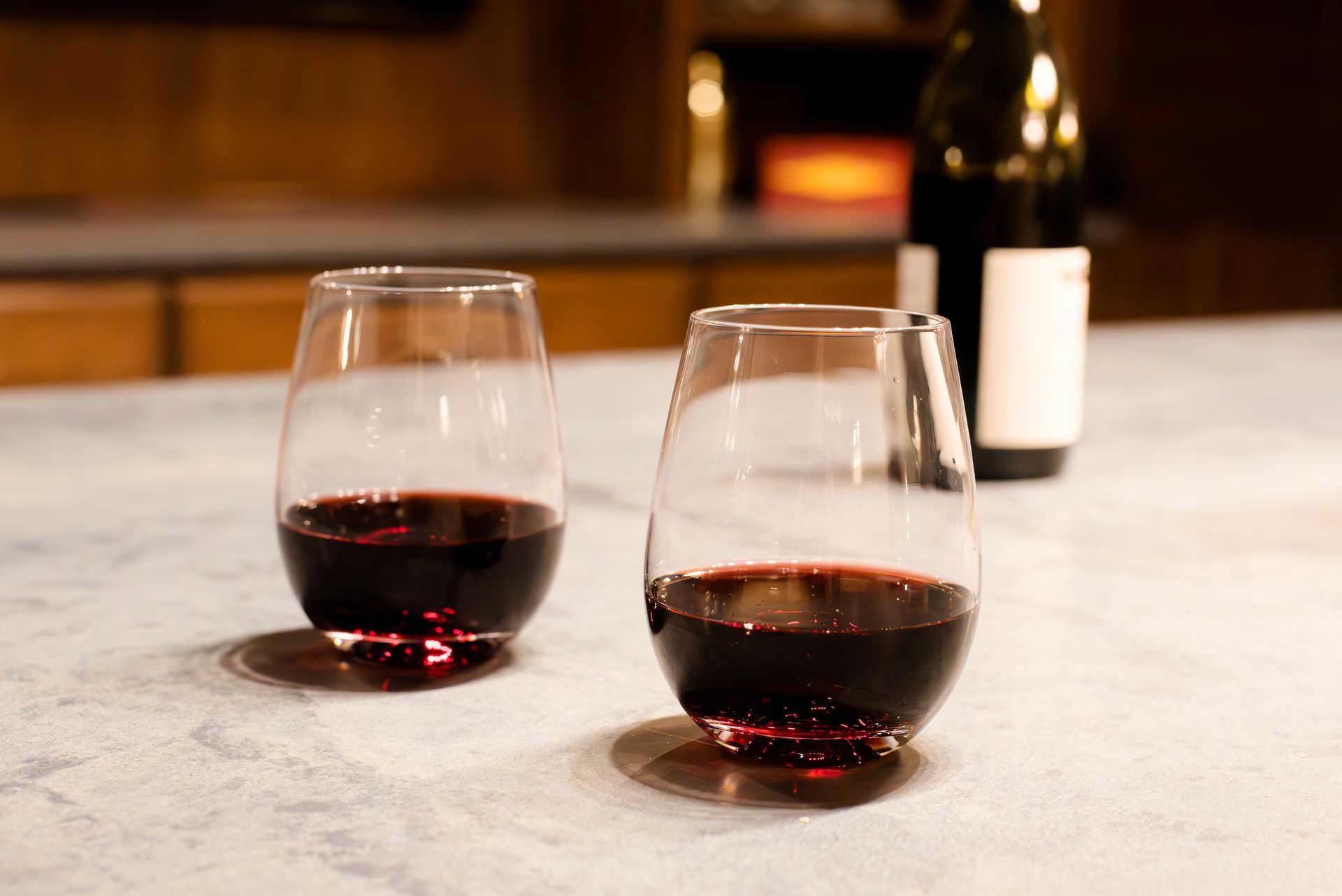 Two stemless wine glasses with red wine and a bottle on a counter.