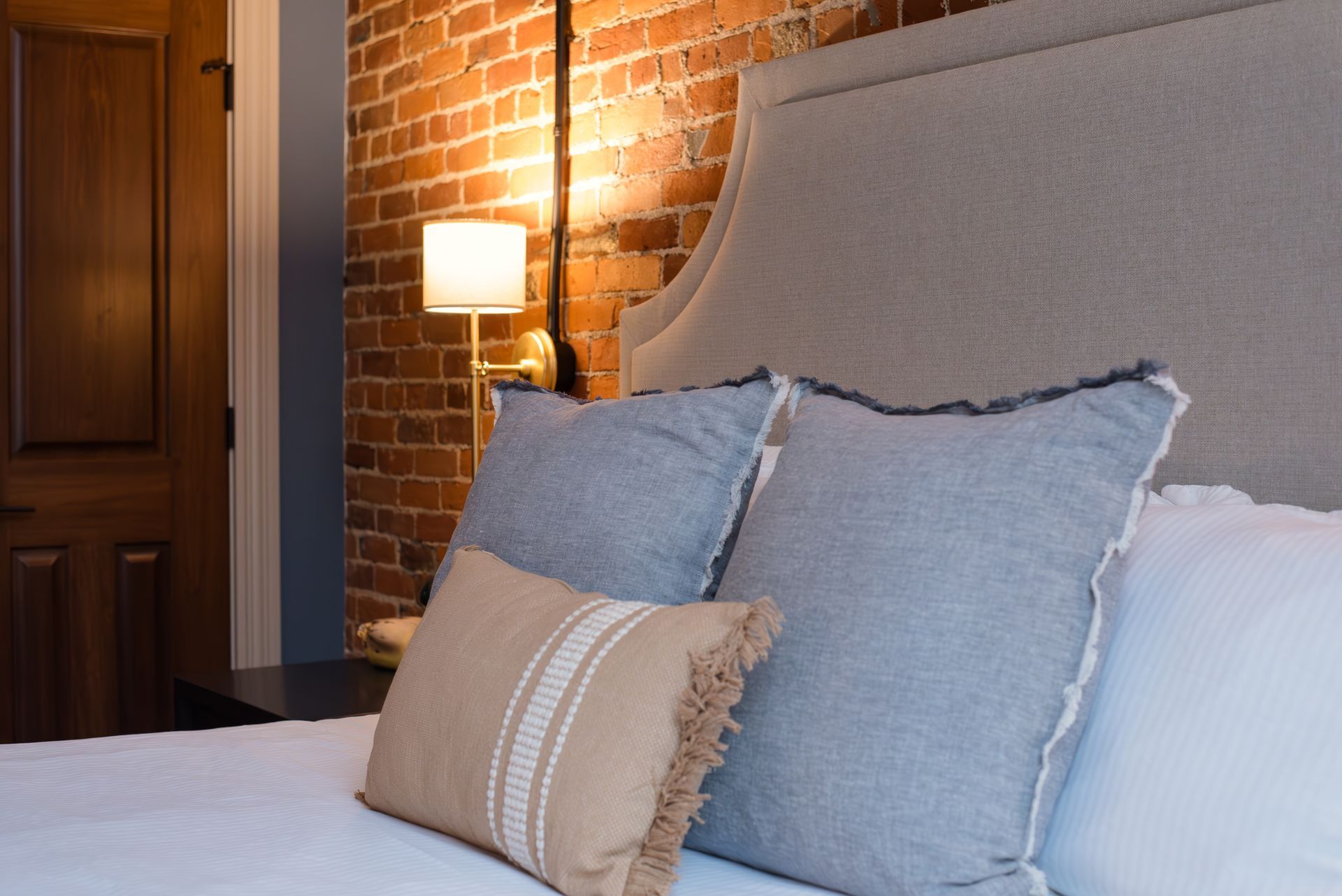 Bedroom with a brick wall, bed with gray headboard and pillows, lamp.