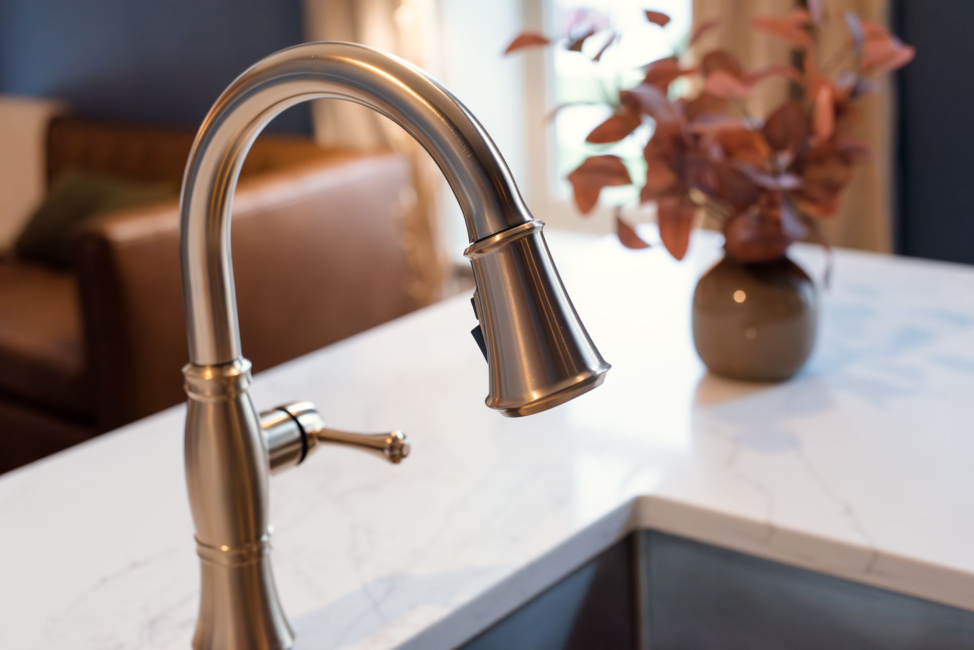 Close-up of a gold kitchen faucet on a white countertop, brown leather chair in the background.