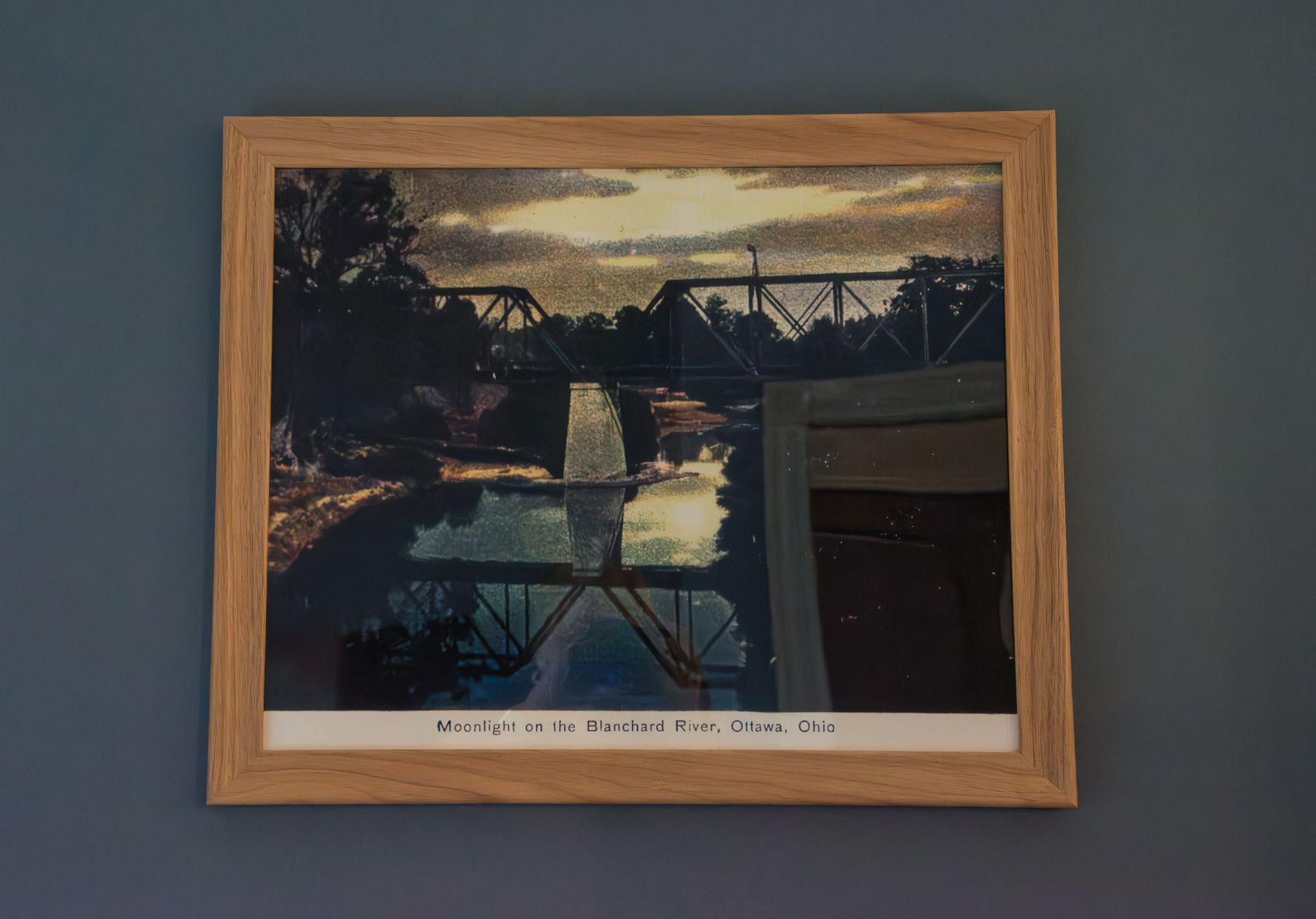Photo of a metal bridge over a river, reflecting in the water. Cloudy sky, framed, hung on a blue wall.