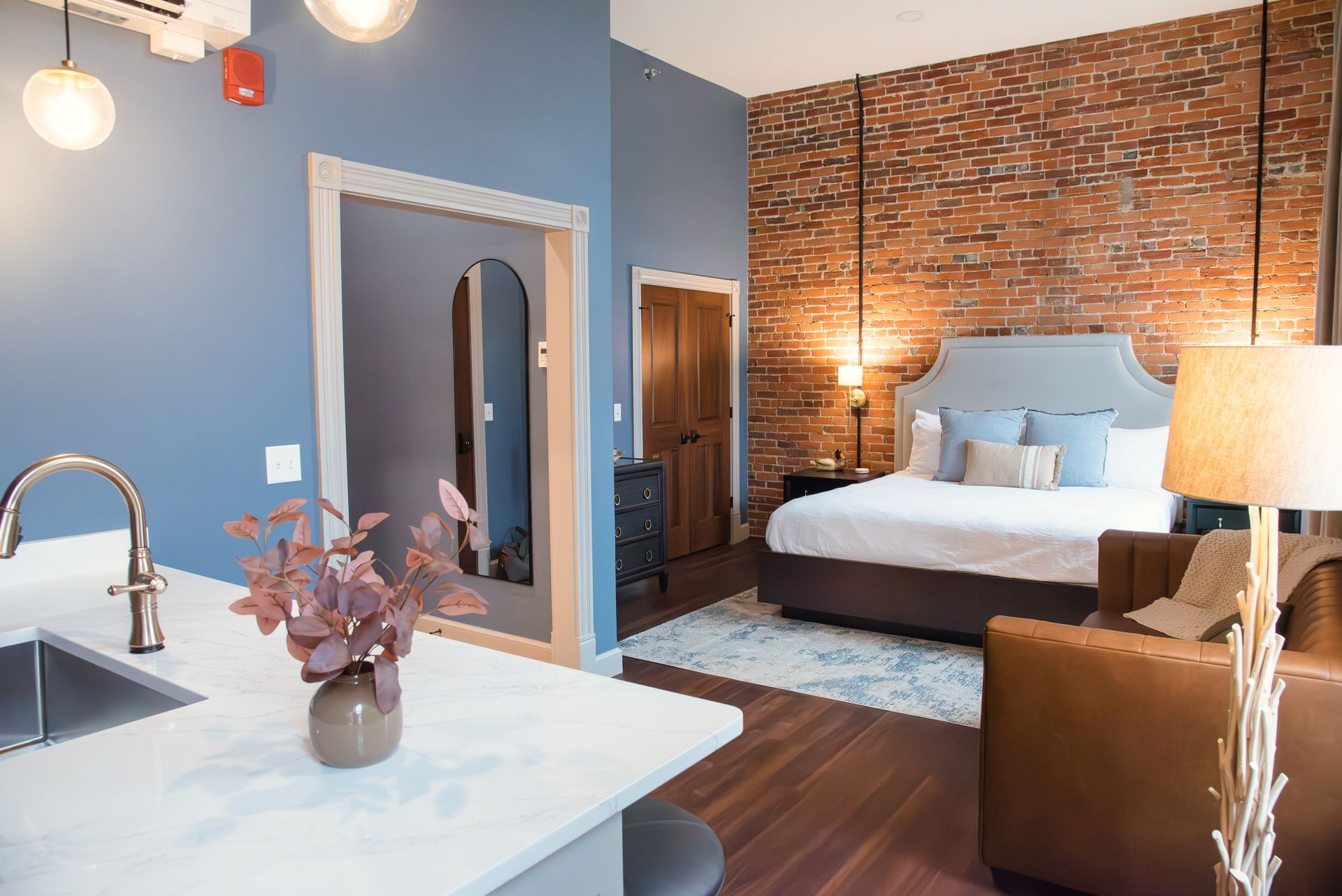 A modern hotel room with a bed against a brick wall, kitchenette, and a leather armchair.