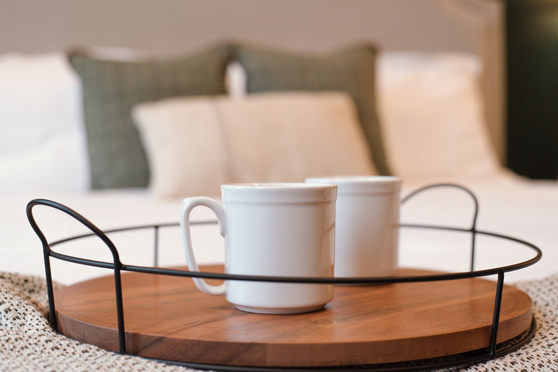 Two white mugs on a wooden tray with black handles, set on a bed with pillows.