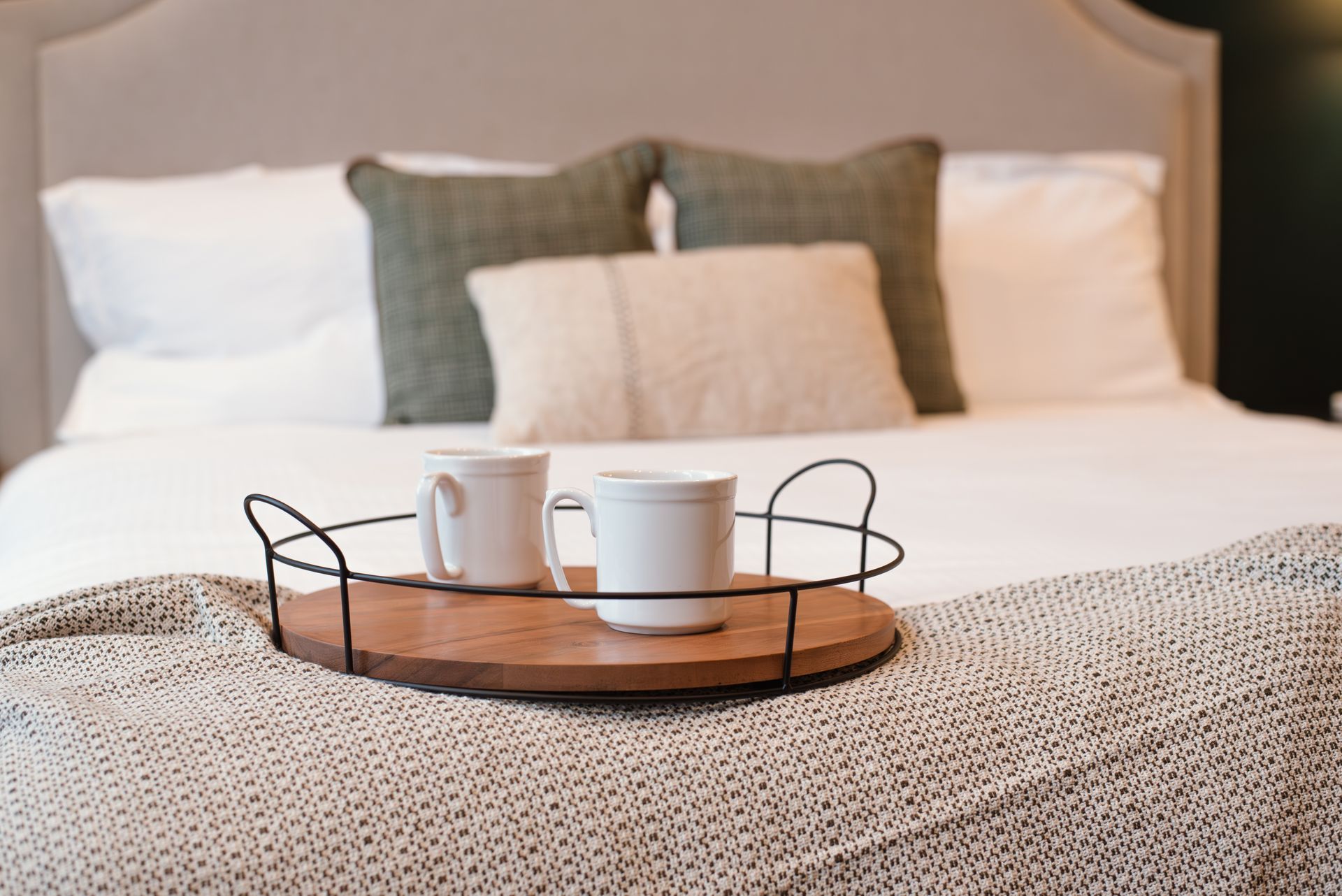 Two white mugs on a wooden tray, on a bed with pillows; soft, neutral tones.
