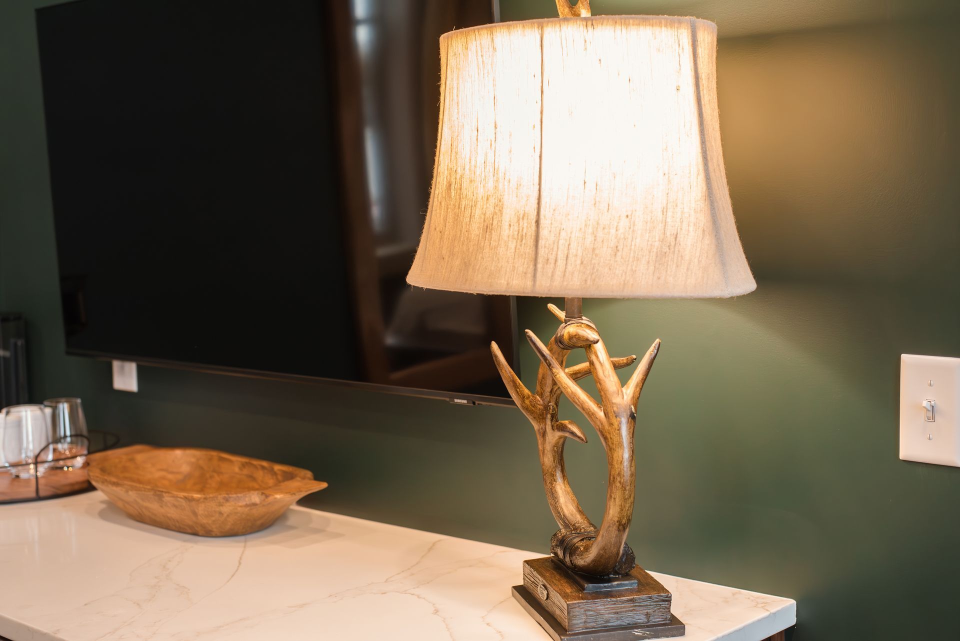 A table lamp with an antler base sits on a white surface. Green wall and TV are in the background.
