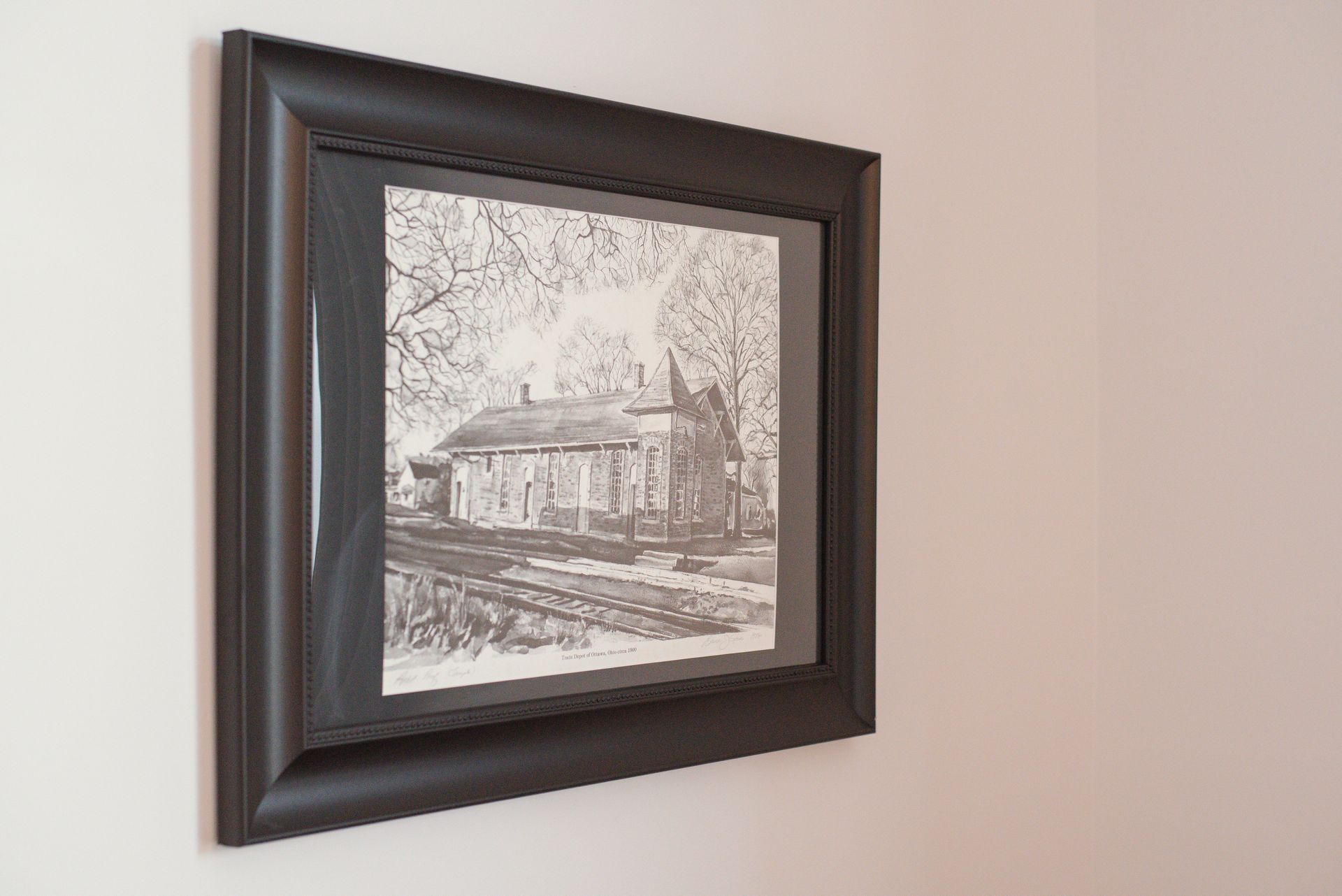 Black framed artwork on a wall, depicting a grayscale drawing of a building and train tracks.