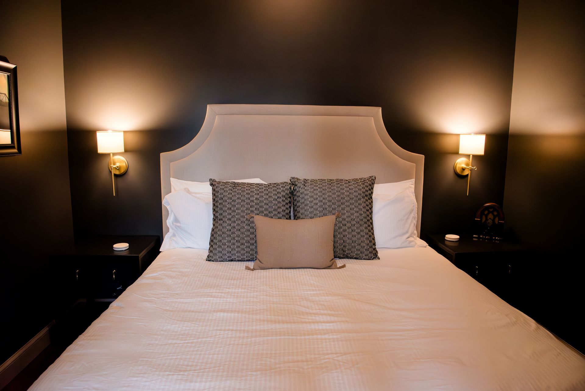 A bed in a dark room with neutral-toned bedding, headboard, and decorative pillows, flanked by bedside tables and sconces.
