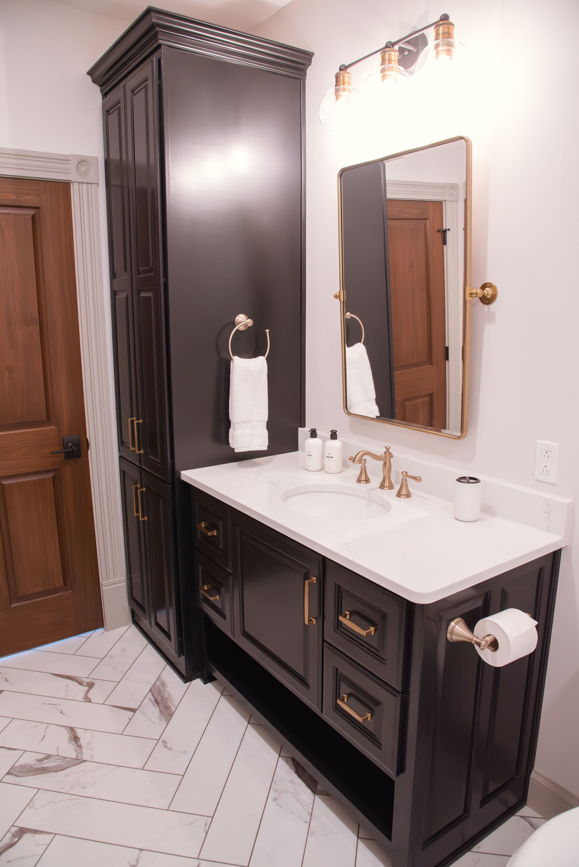 Black vanity with gold hardware, white countertop, and matching tall storage cabinet in a modern bathroom.