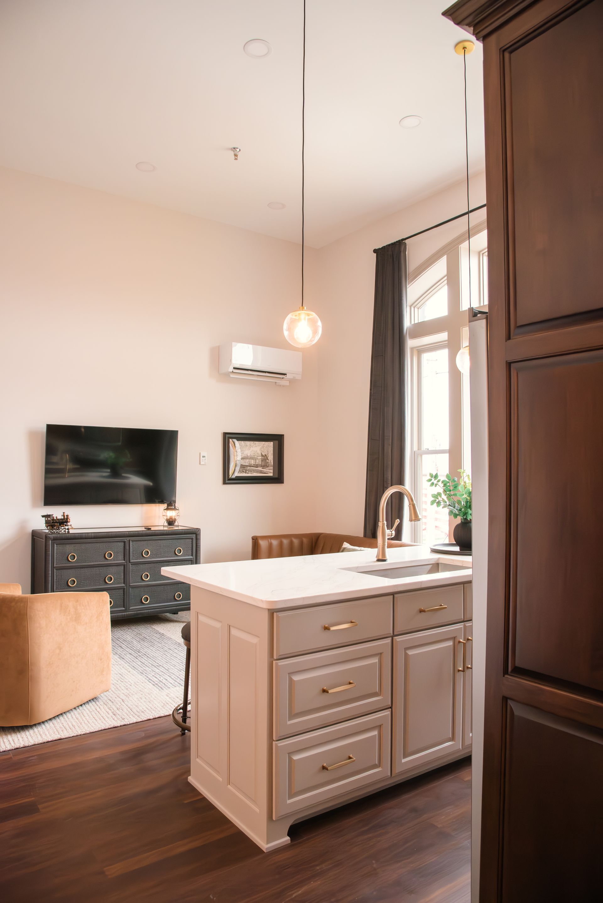 Kitchen island with light fixture, TV, and window. Beige cabinets, gold accents, and dark wood floor.