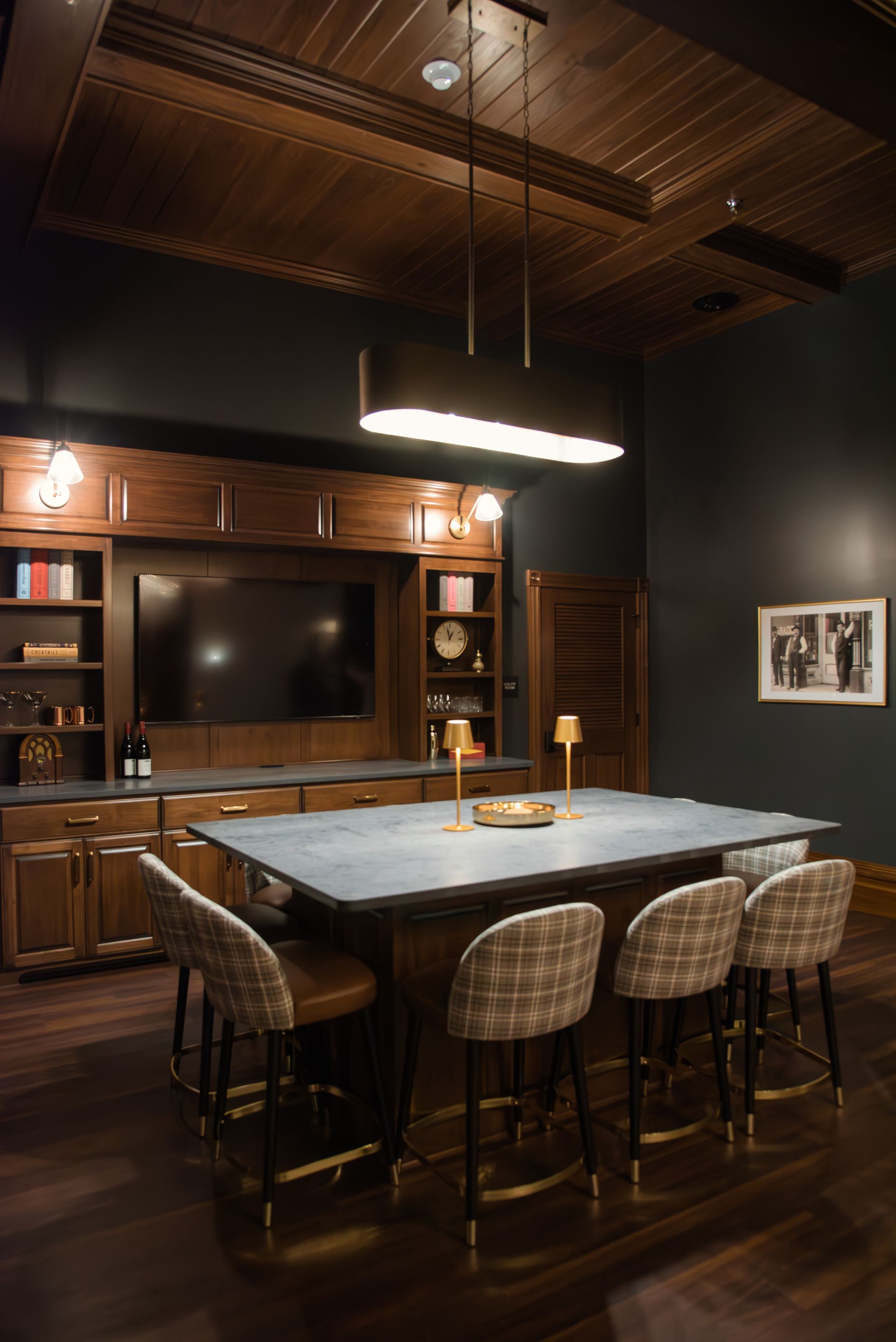 Dark-toned bar with a large table, chairs, and TV. The space has dark wood, dim lighting, and a sophisticated feel.
