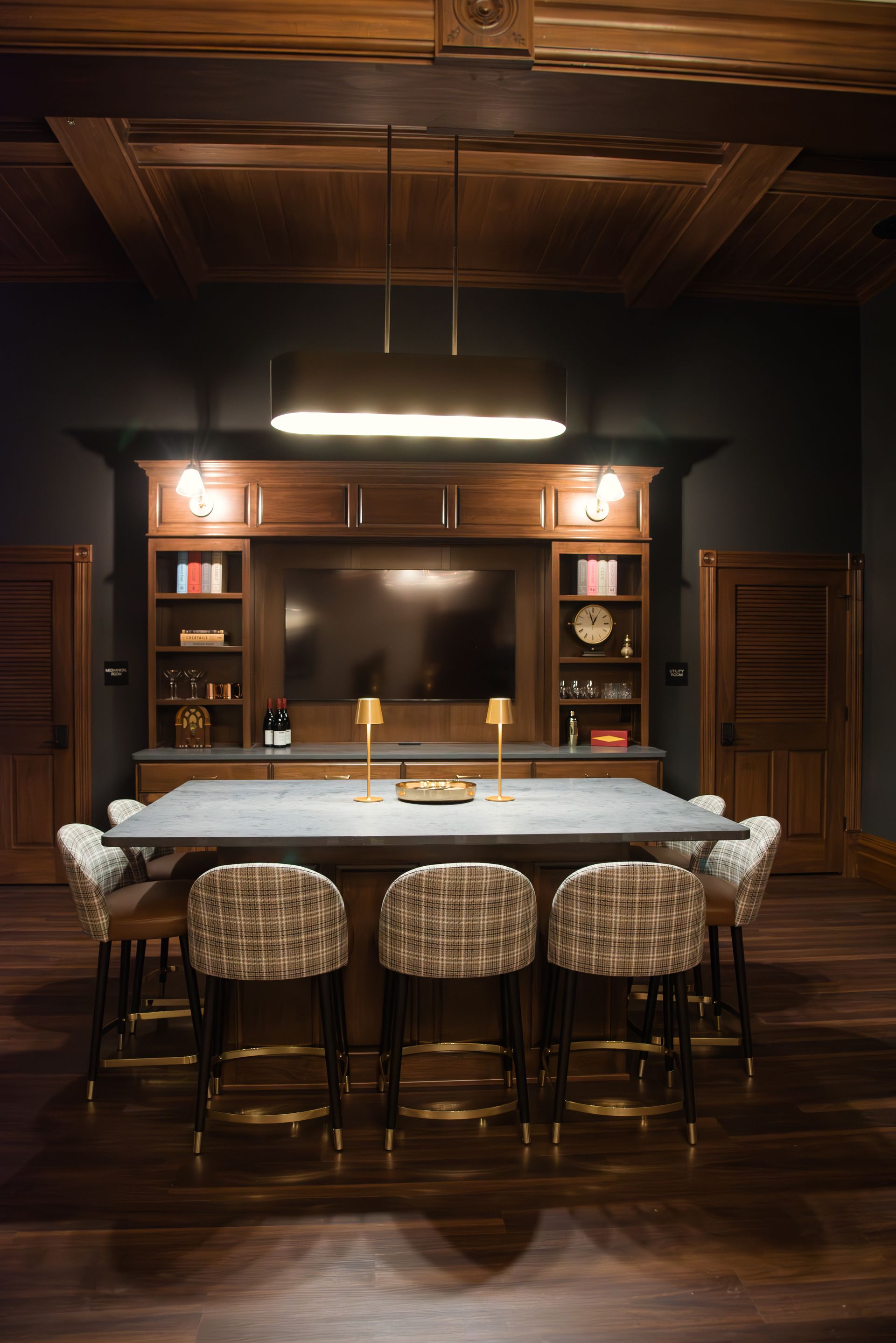 Dark wood bar with stools, TV, shelves, and overhead lighting in a room with dark walls and wood flooring.