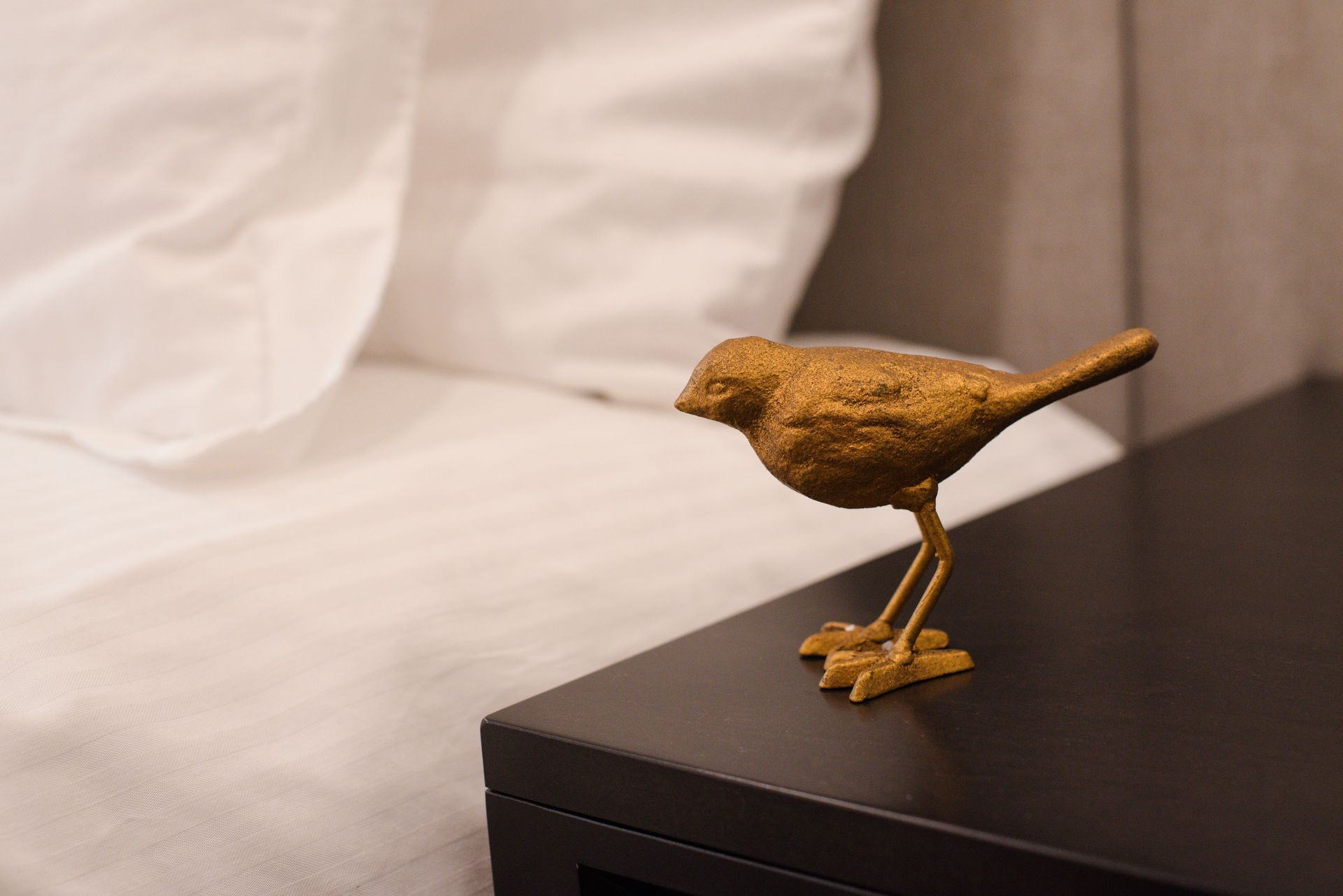 Golden bird figurine on a black nightstand next to a bed with white linens.