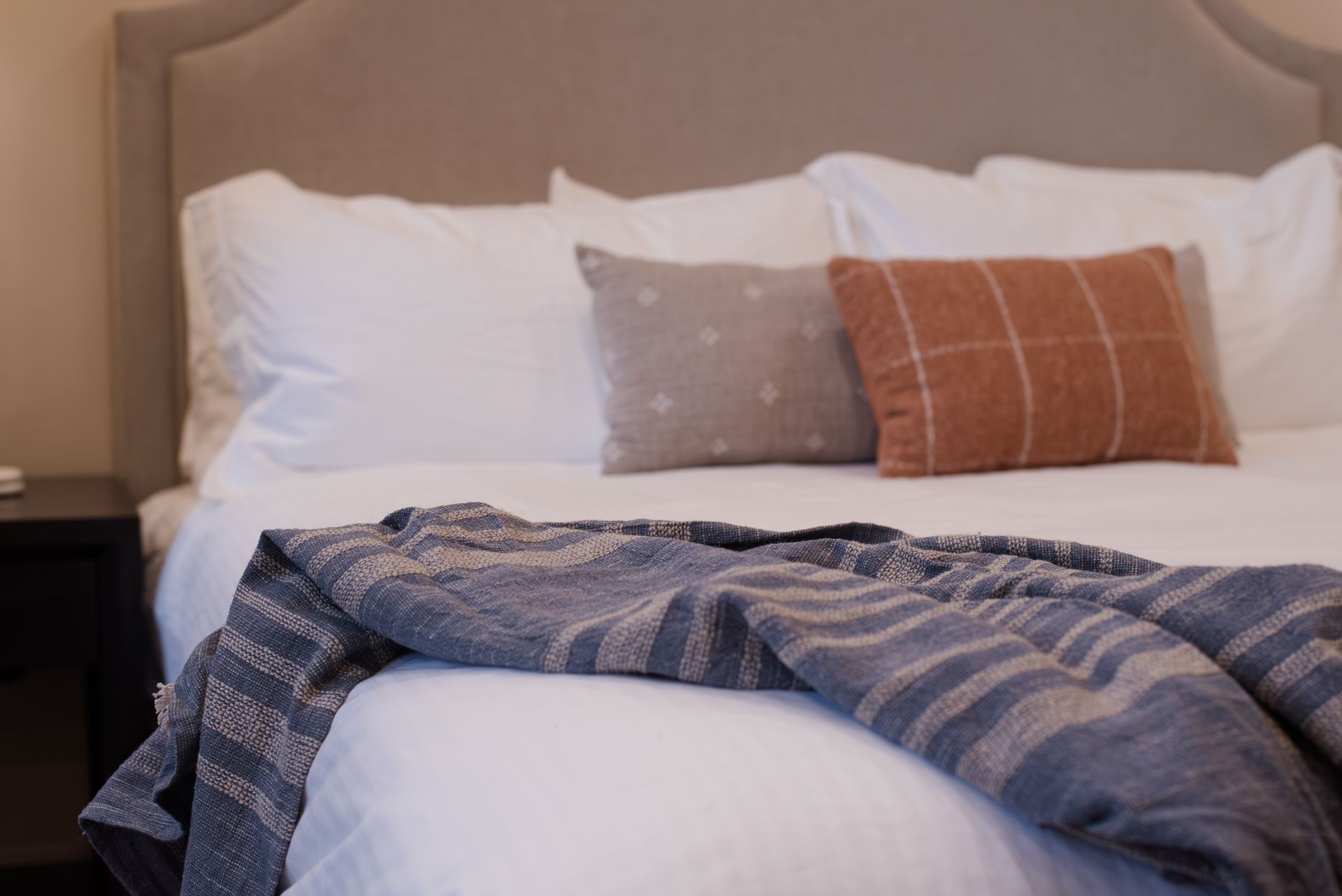 Bedroom bed with white bedding, pillows, and a blue blanket. Two decorative pillows and a nightstand.