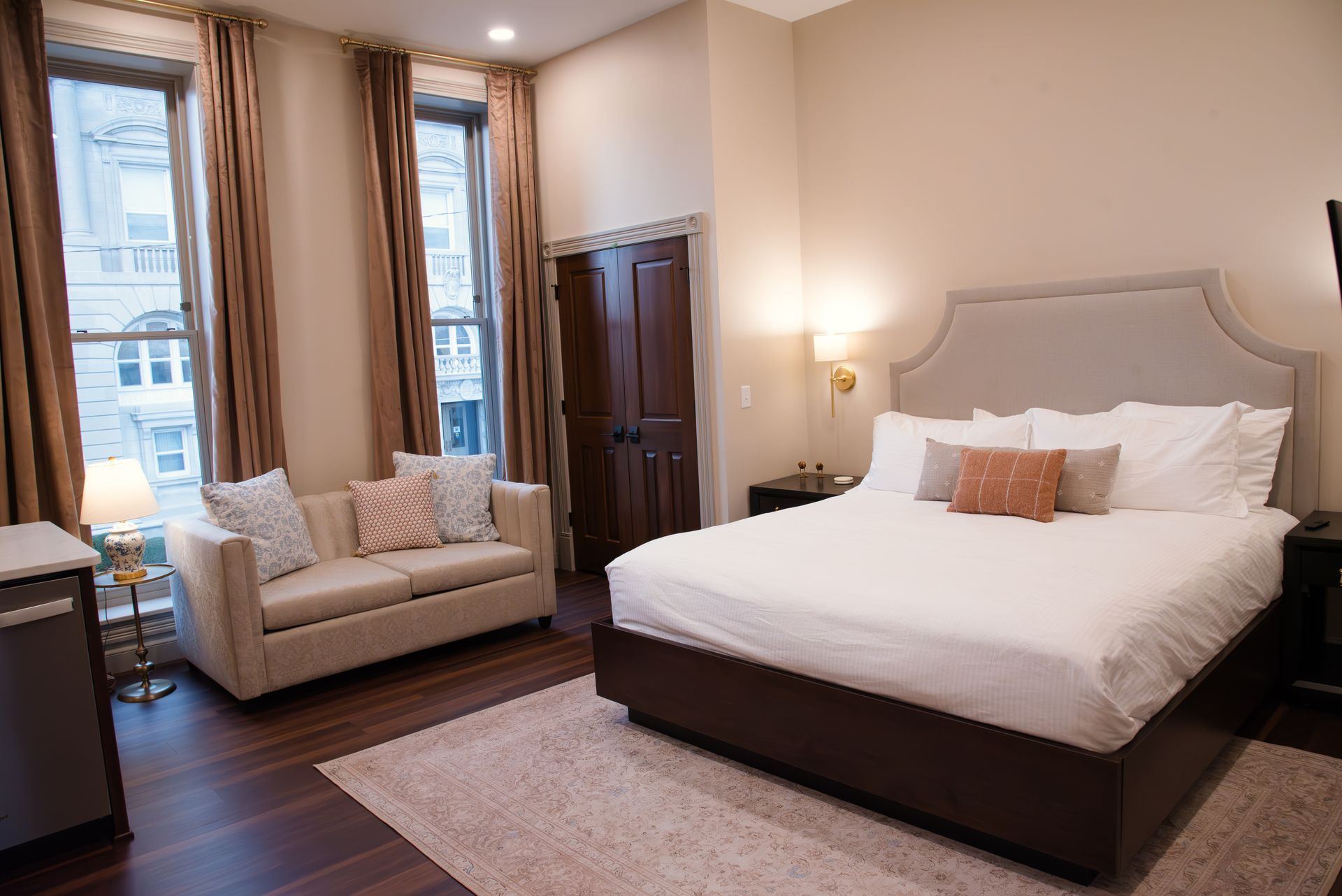 Hotel room with bed, sofa, windows, and dark wood flooring.