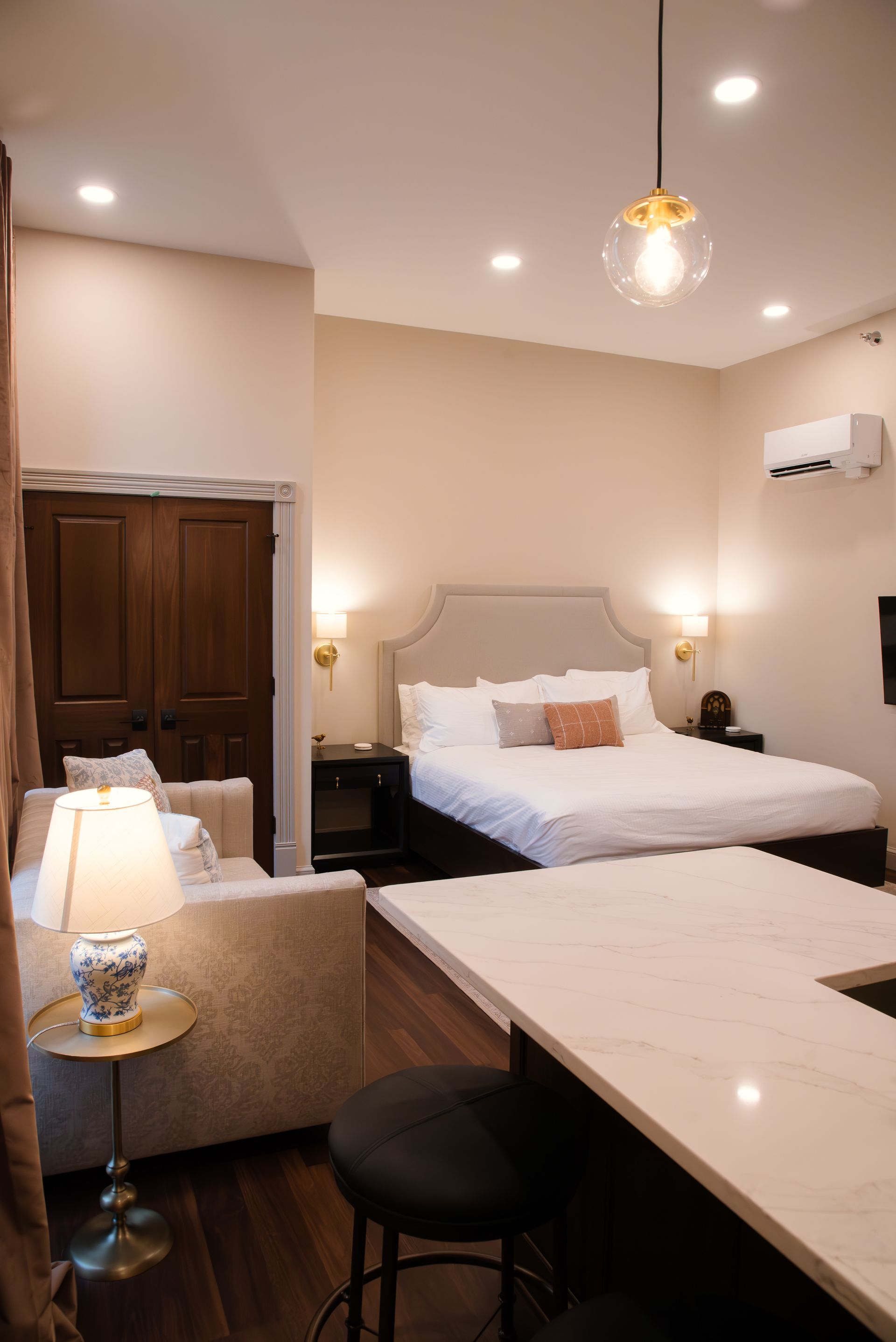 Cozy hotel room with a bed, seating area, and kitchen bar; neutral colors, warm lighting.