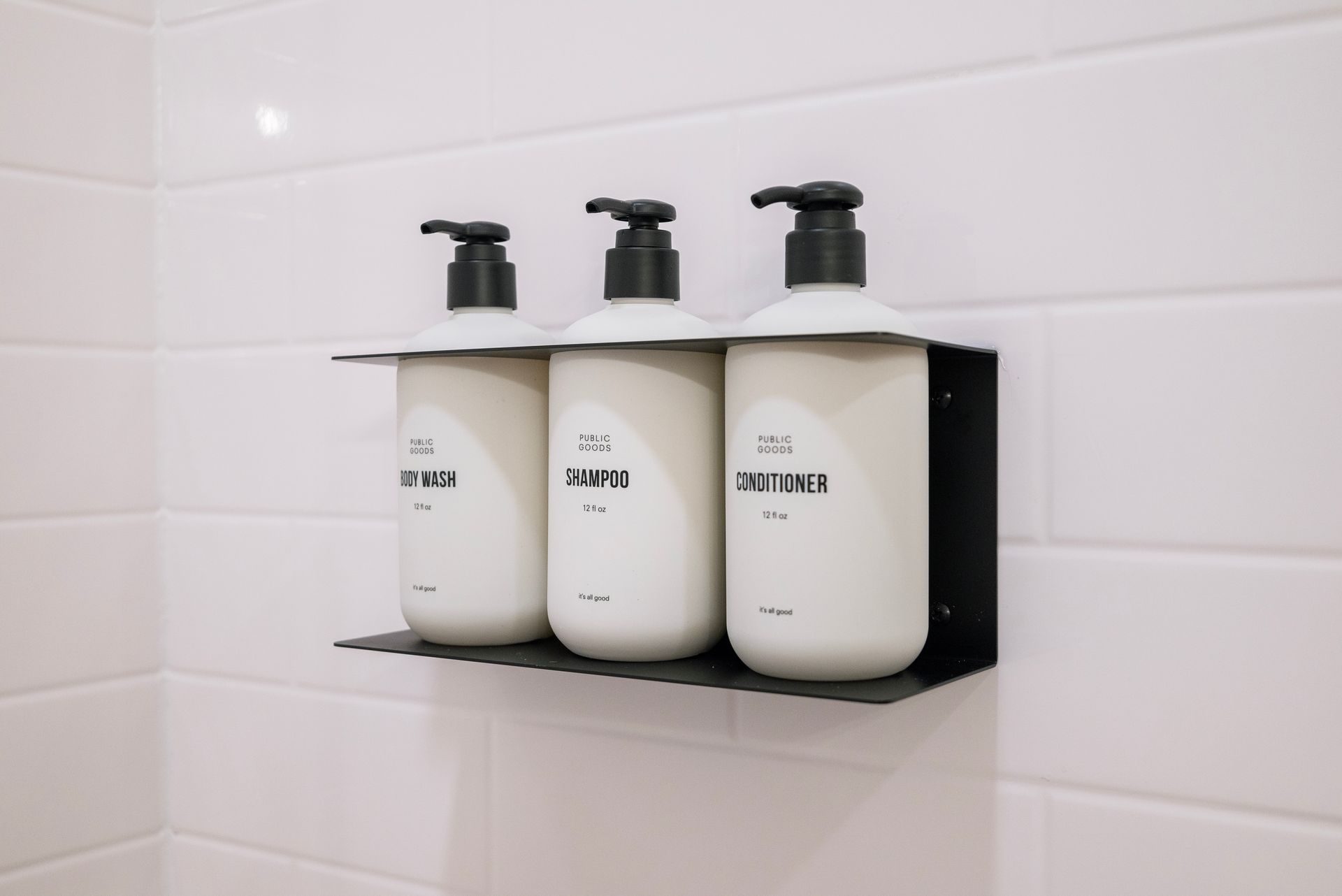 Three white shampoo bottles with black pumps on a black shelf, mounted on white tiled shower wall.