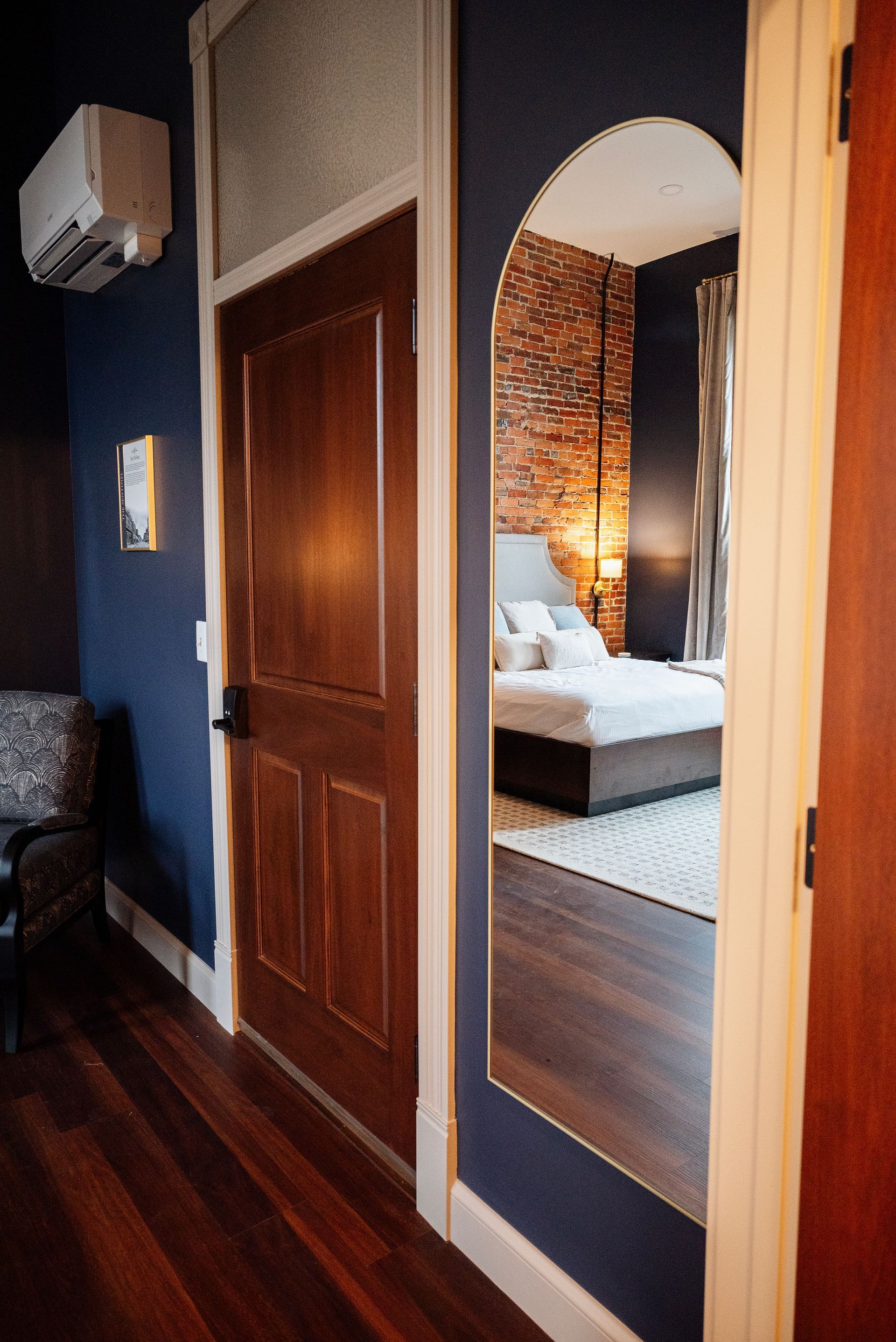 Hallway with wooden door and arched doorway revealing a bedroom with brick wall and bed.