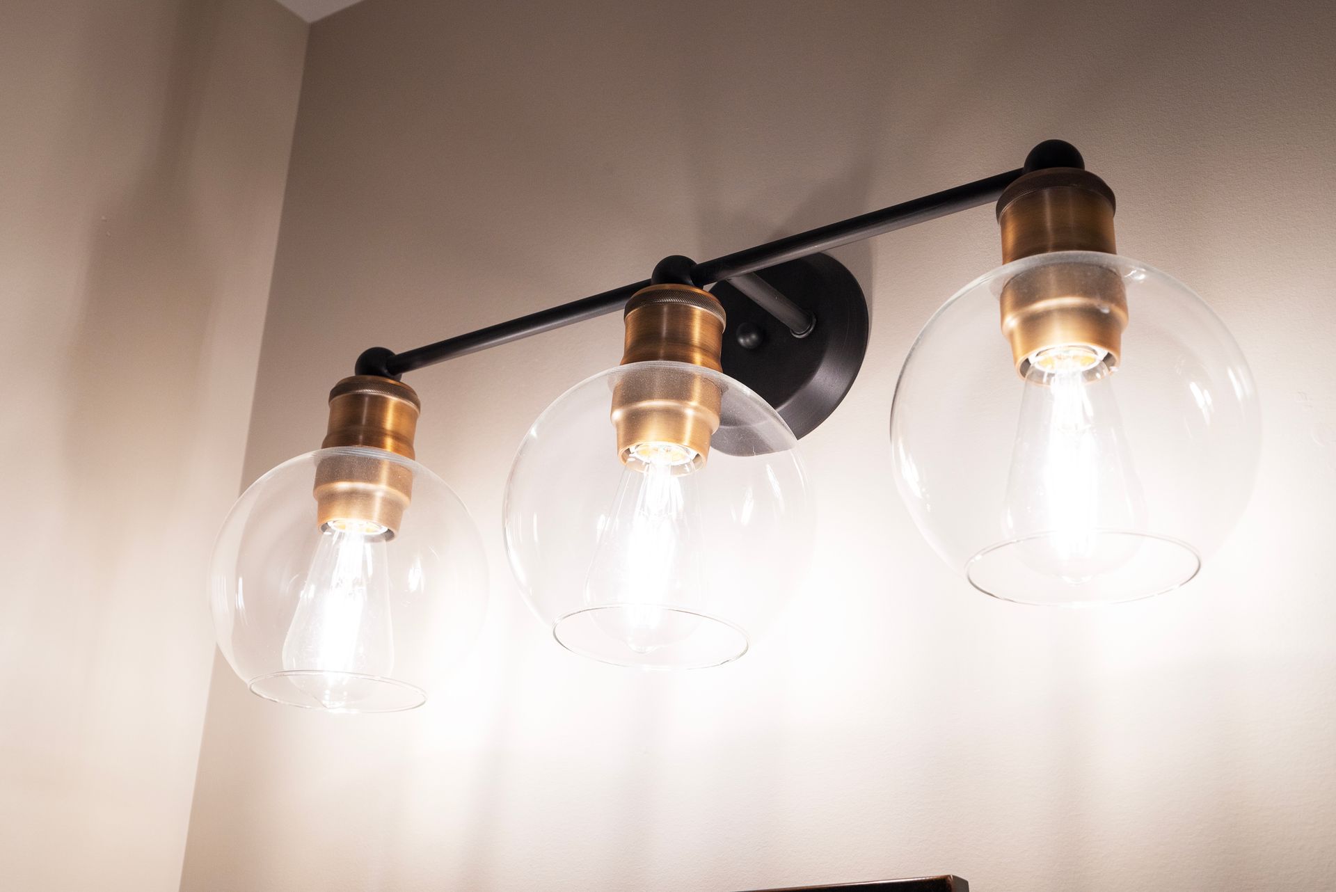 Three-light vanity fixture with clear globe shades and brass-colored accents, mounted on a light-colored wall.