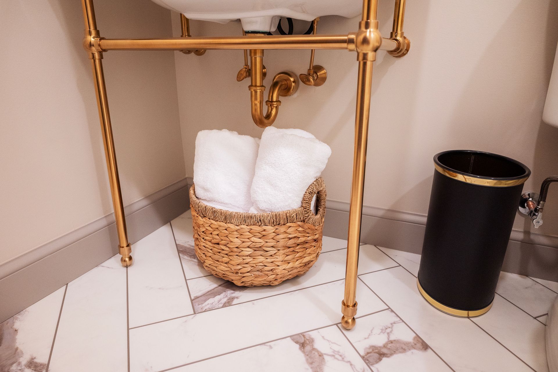 White towels in a wicker basket under a gold-legged sink. Black trash can with gold trim beside.