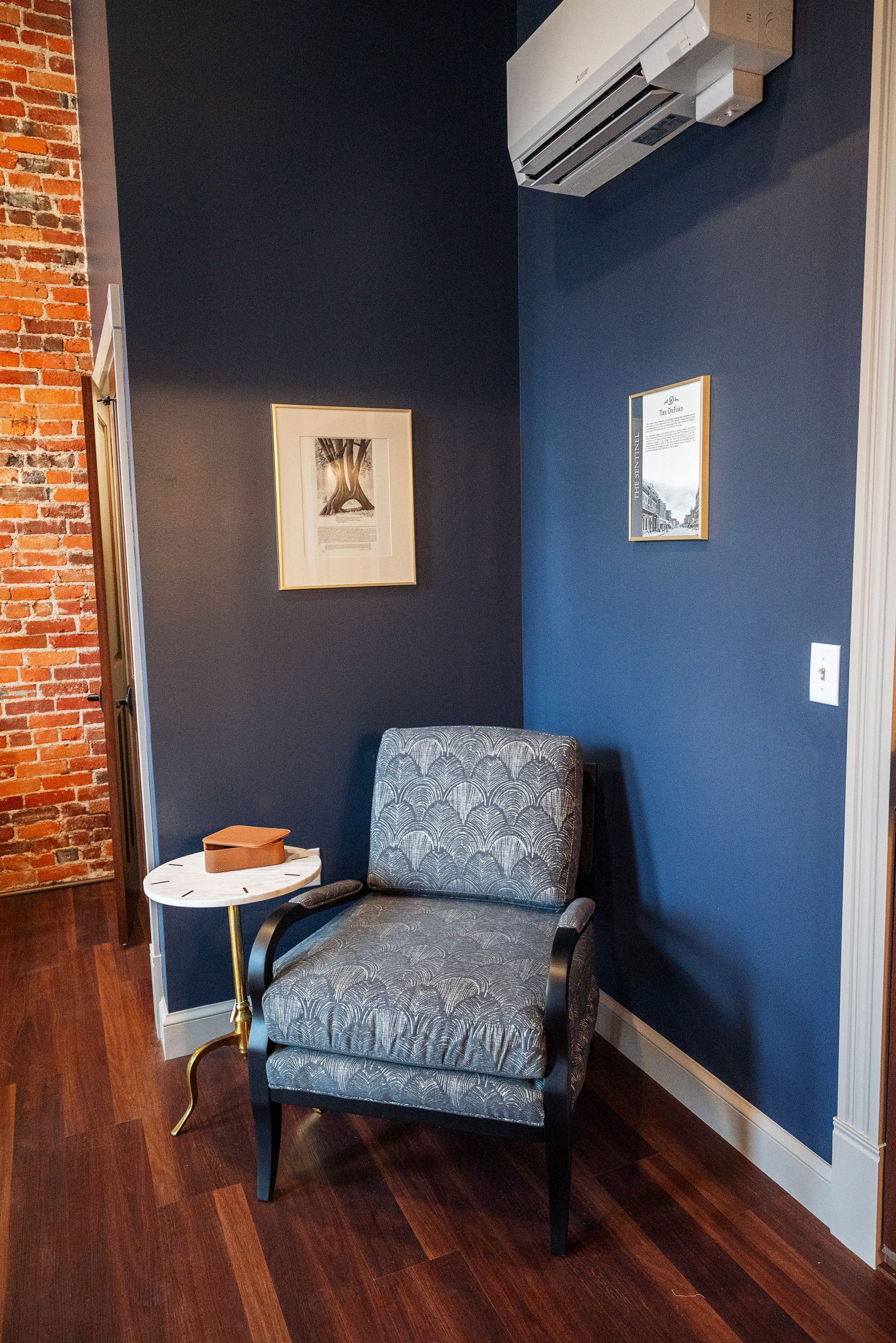 Cozy corner with patterned armchair, small side table, blue textured walls, brick, and two framed prints.