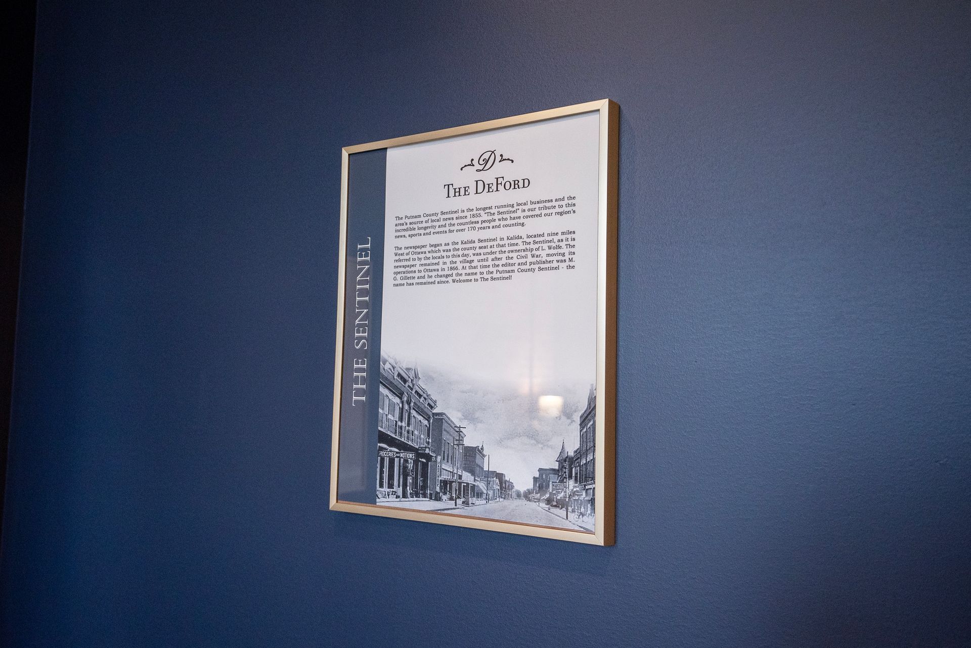 Framed artwork on a dark blue wall. The piece shows a town street scene and text, in a gold frame.