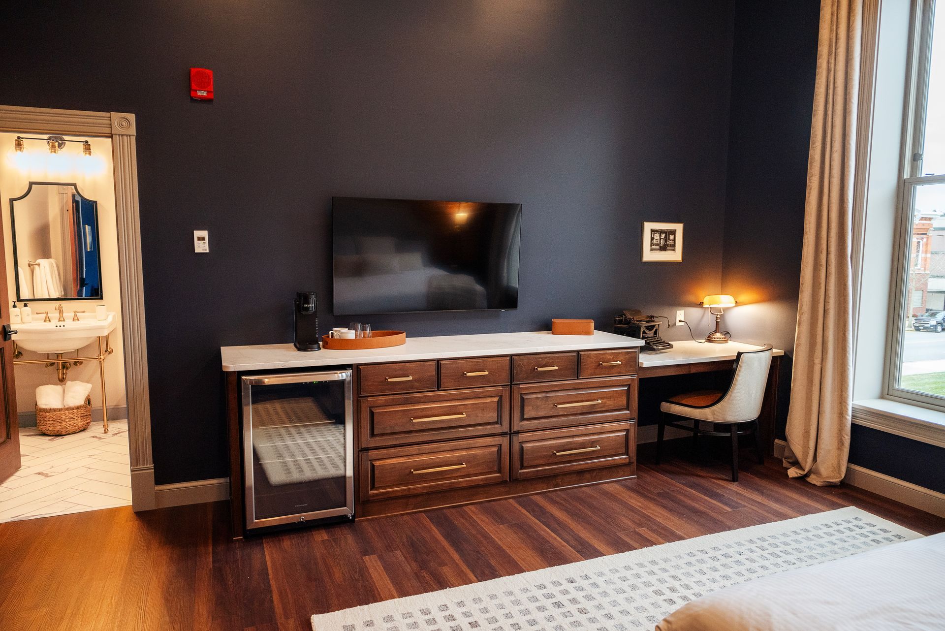 Hotel room with dark blue wall, TV, wooden dresser, desk, and an open bathroom.