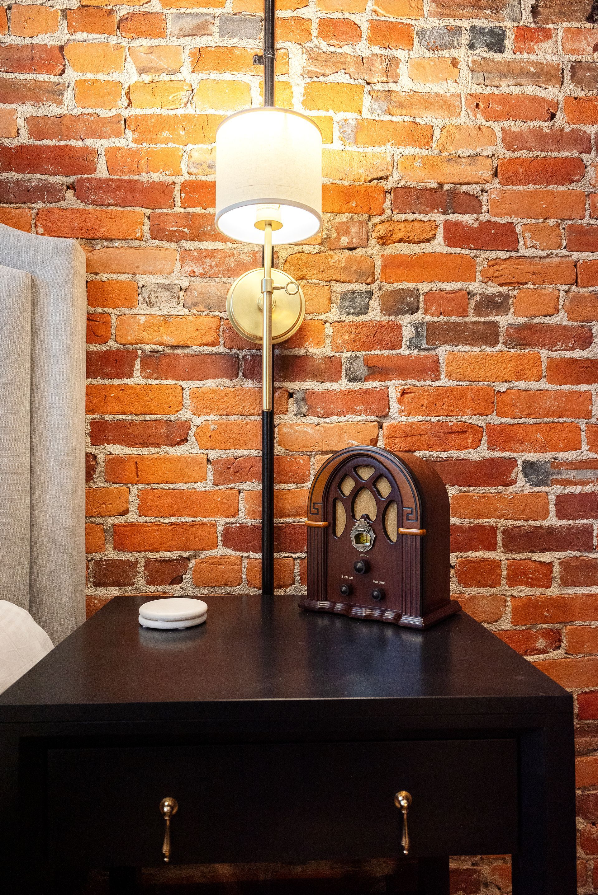 Black nightstand with lamp against a red brick wall.  A radio and a small white object are on the nightstand.