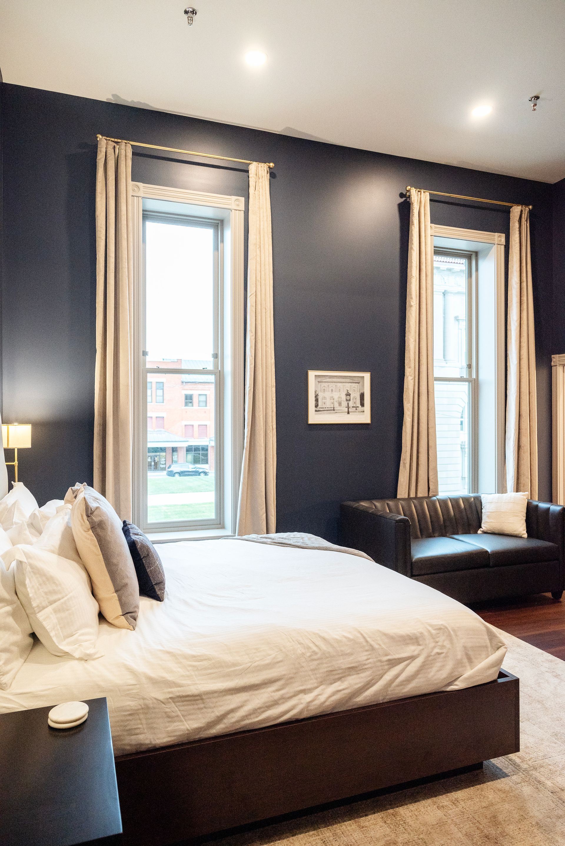 Bedroom with dark blue walls, large bed, two windows, and a dark leather sofa.
