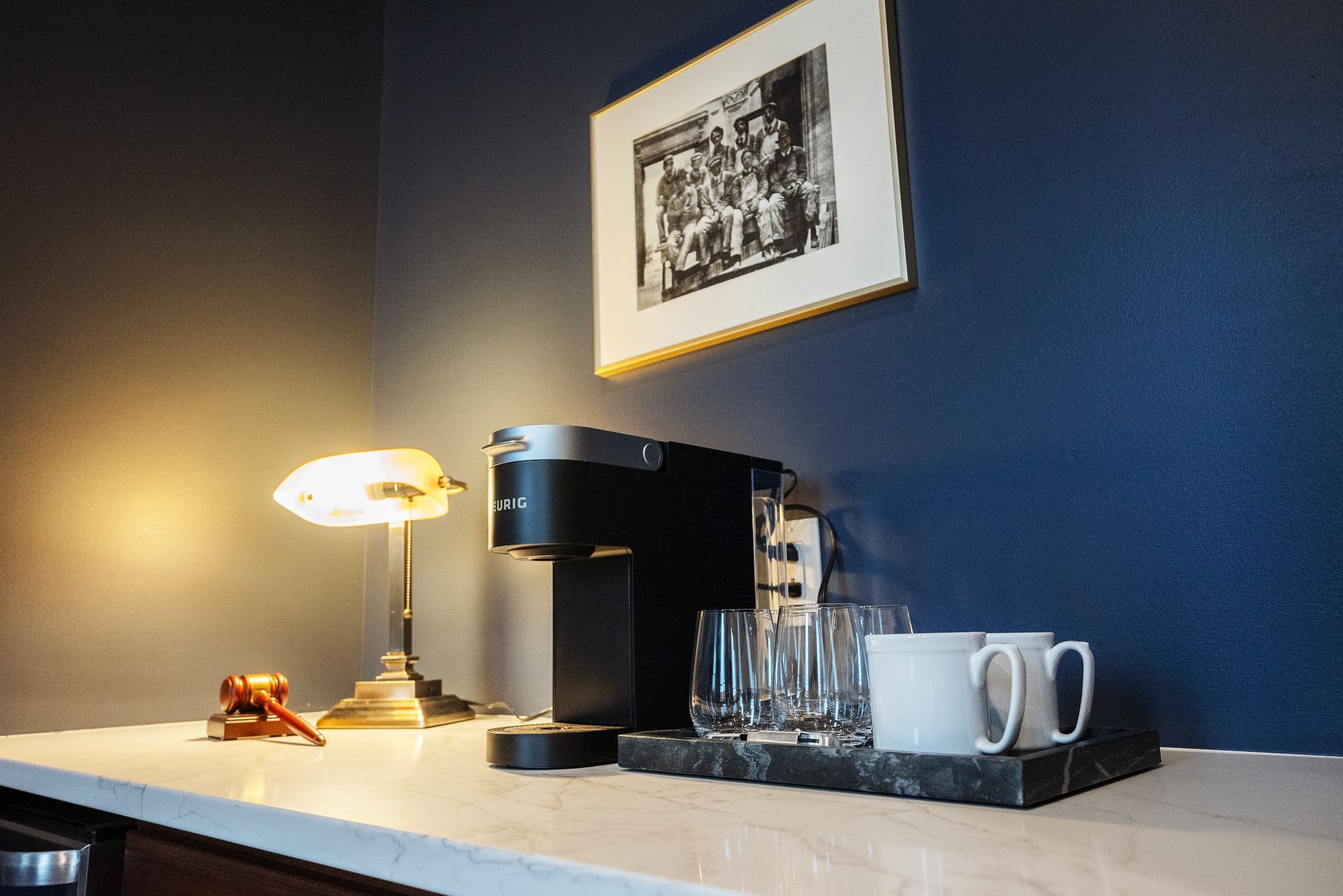Coffee station with Keurig, mugs, and glasses on a marble tray under a lamp, art on a blue wall.