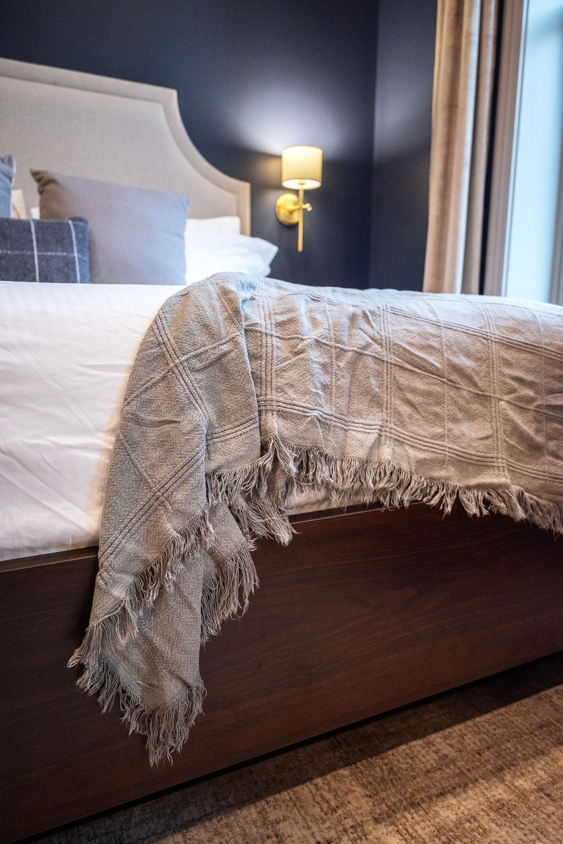 Bed with silver fringed blanket, beige headboard, and sconce on dark blue wall.