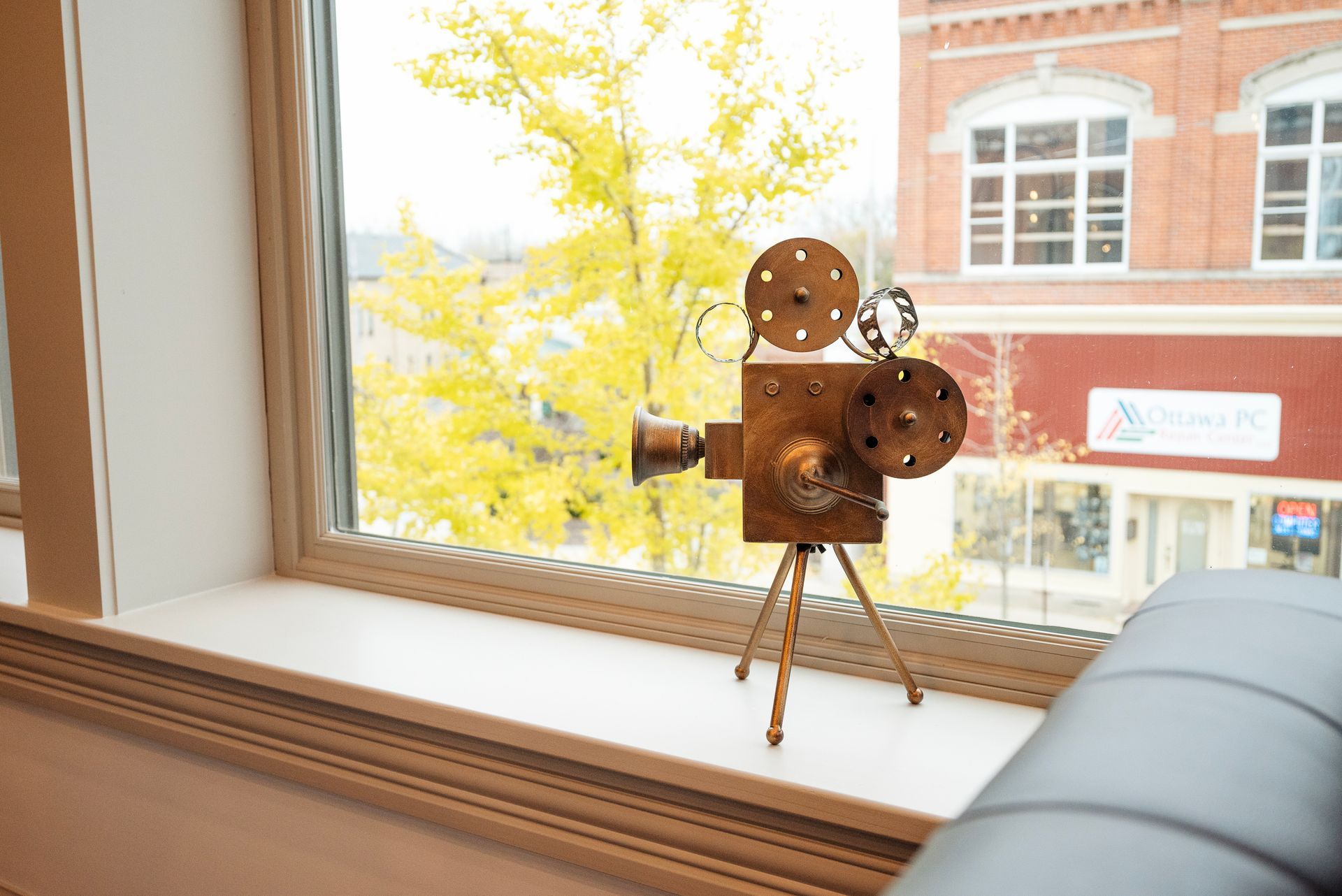 Decorative metal film camera on a windowsill, framed by a yellow tree and a brick building.