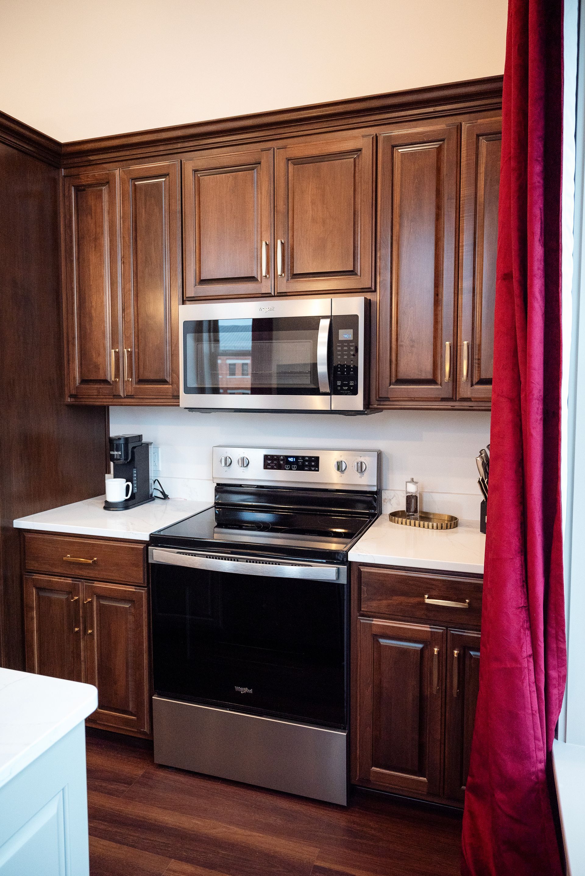 Brown kitchen cabinets with a stainless steel oven and microwave. White countertops and a red curtain are visible.