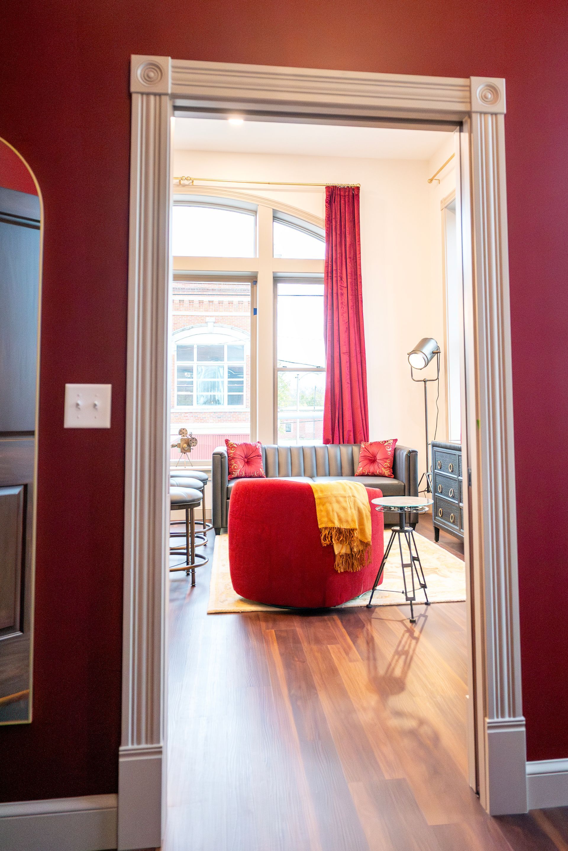 Looking through a doorframe into a living room with red ottoman, couch, and curtain.