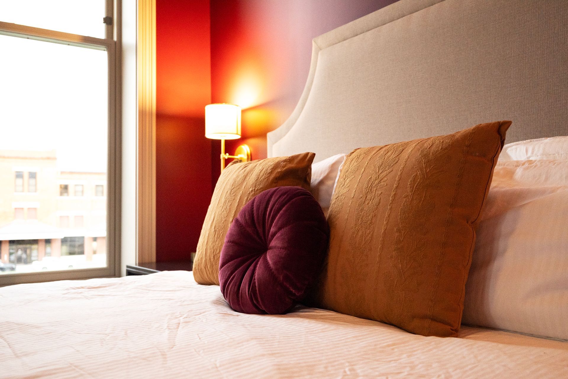 Bed with pillows in front of a window, room with red wall, lit lamp on the background.