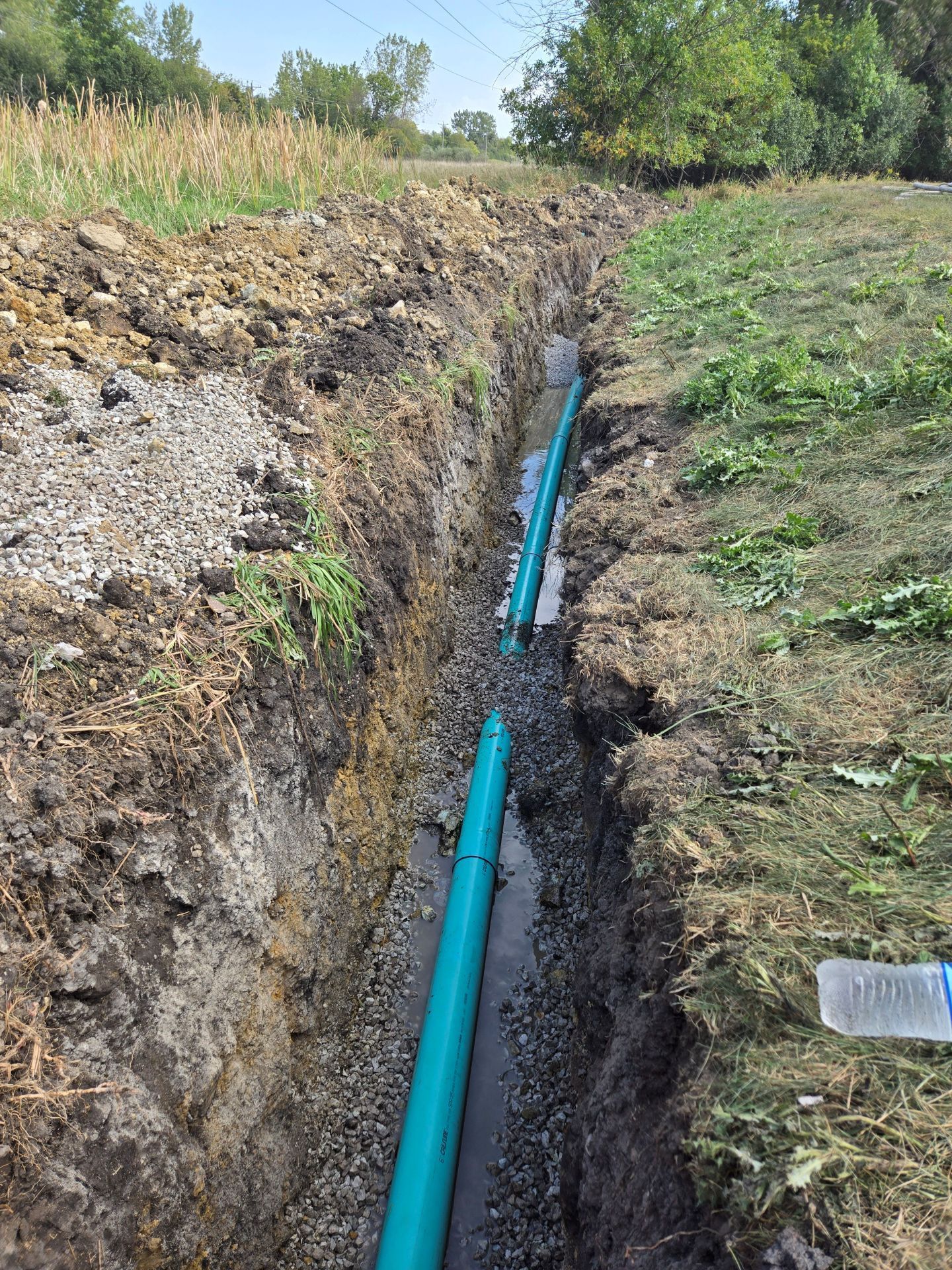 Trench with blue pipes installed, gravel base, grassy edges, and a water bottle.
