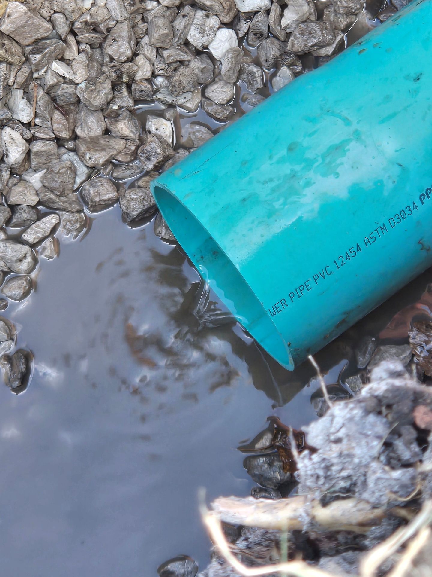 A turquoise PVC pipe drains water into a puddle surrounded by gravel.